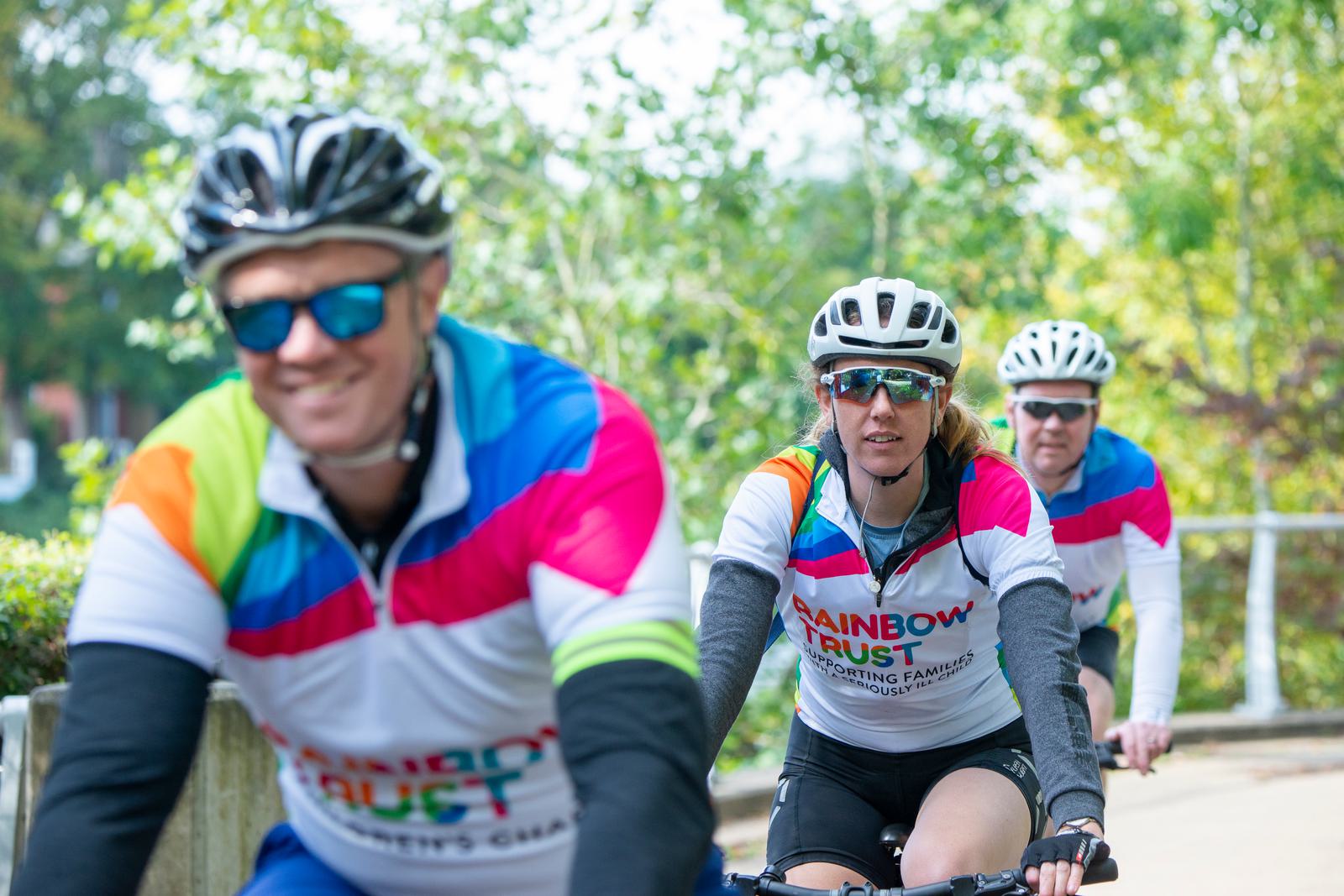 Three cyclists wearing helmets and colorful jerseys ride on a sunny path, lined with green trees. The jerseys have "Rainbow Trust" written on them. The cyclist in the foreground is slightly blurred, while the two behind are in focus.