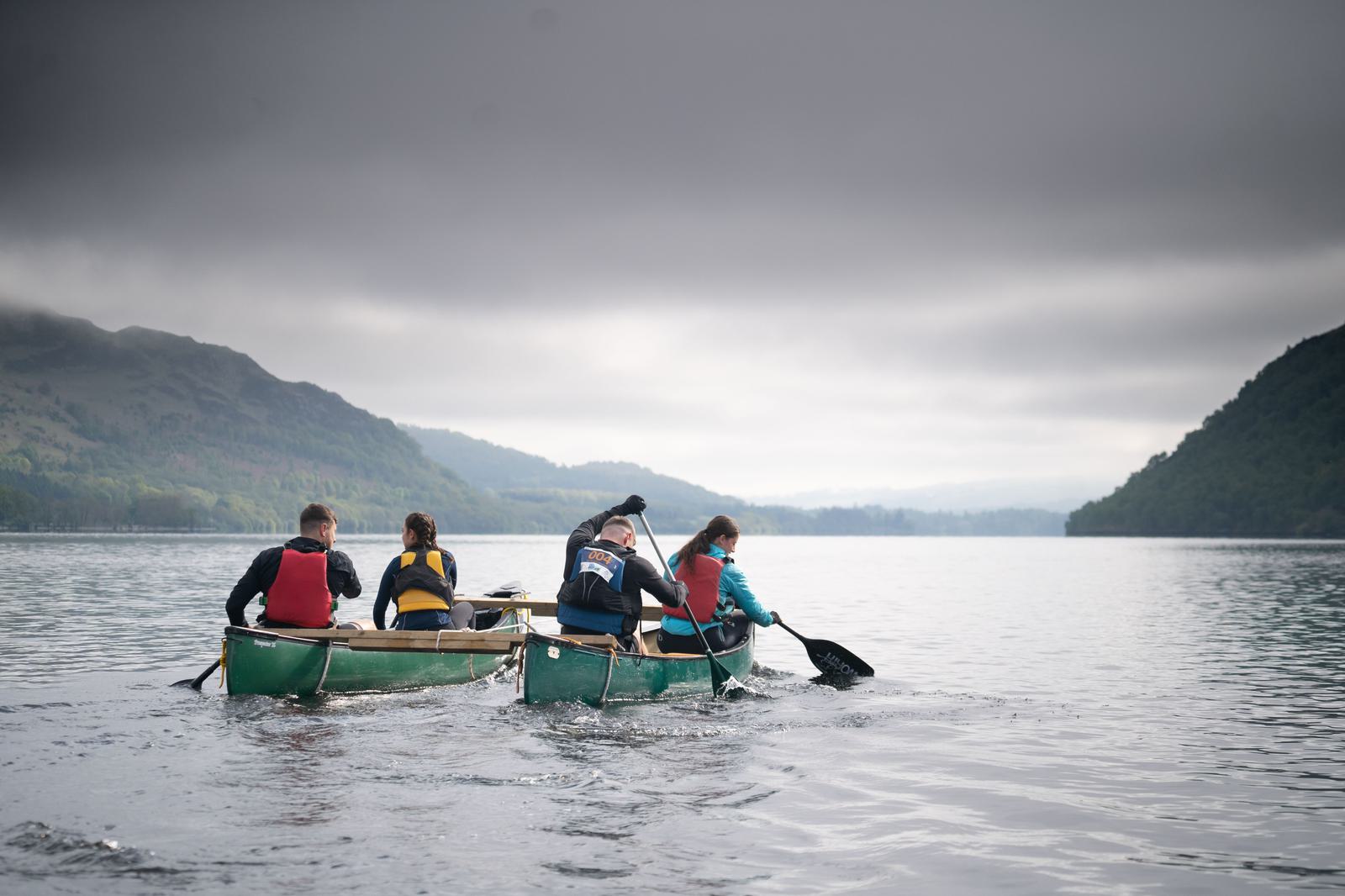Four people paddle two green canoes on a calm lake surrounded by misty mountains under a cloudy sky. The paddlers, wearing life jackets, are seen from behind as they navigate the serene waters together.