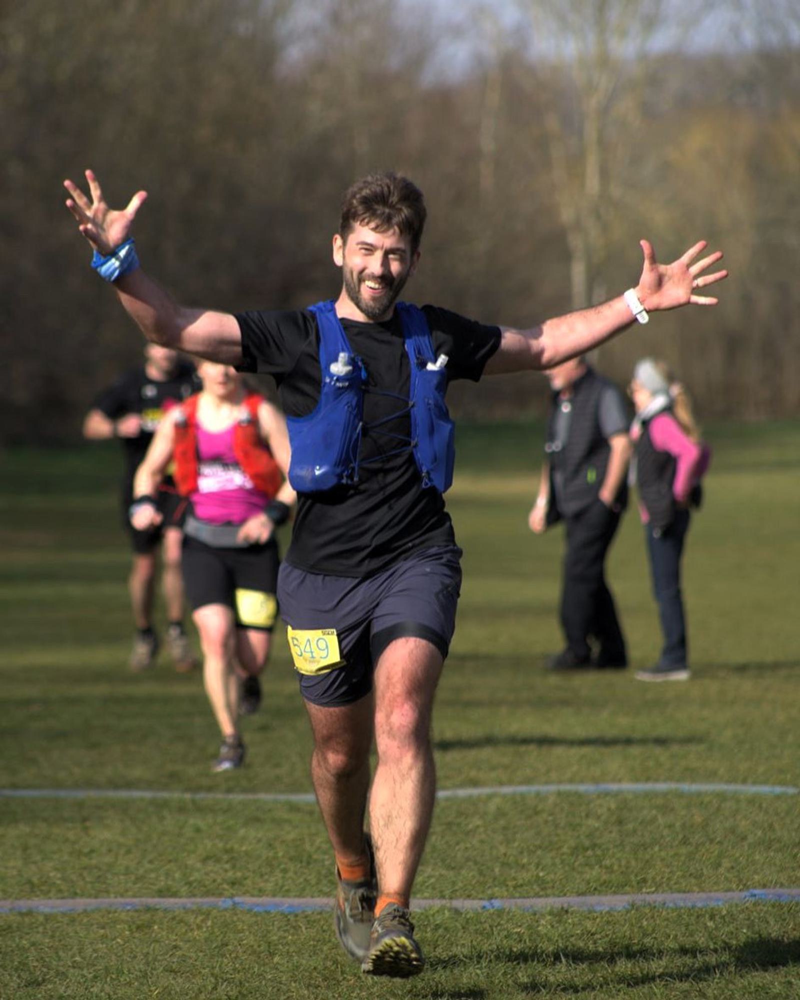 A person in a blue running vest triumphantly crosses a race finish line with arms outstretched. Other runners and spectators are visible in the background, on a grassy area with trees. The sun casts shadows on the ground.