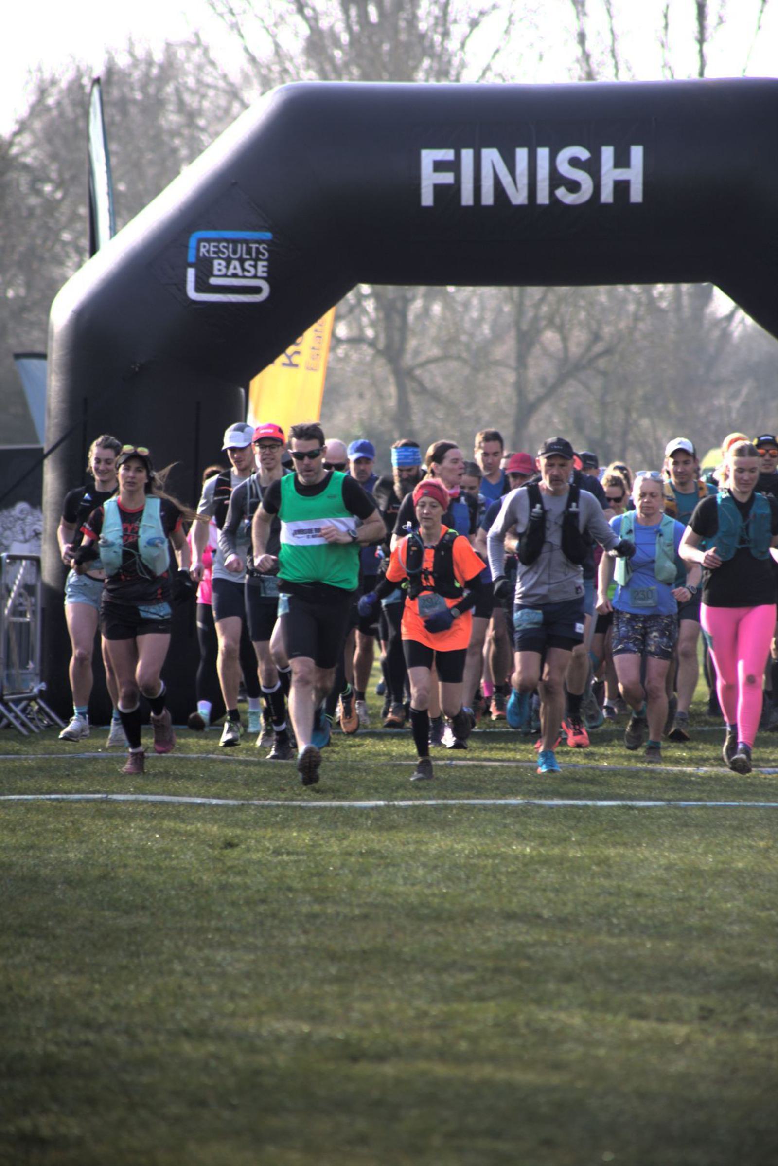 A group of runners in athletic gear approach the finish line during a race. They are outdoors on a grassy field beneath an archway that reads "FINISH." The sky is clear and trees are visible in the background.