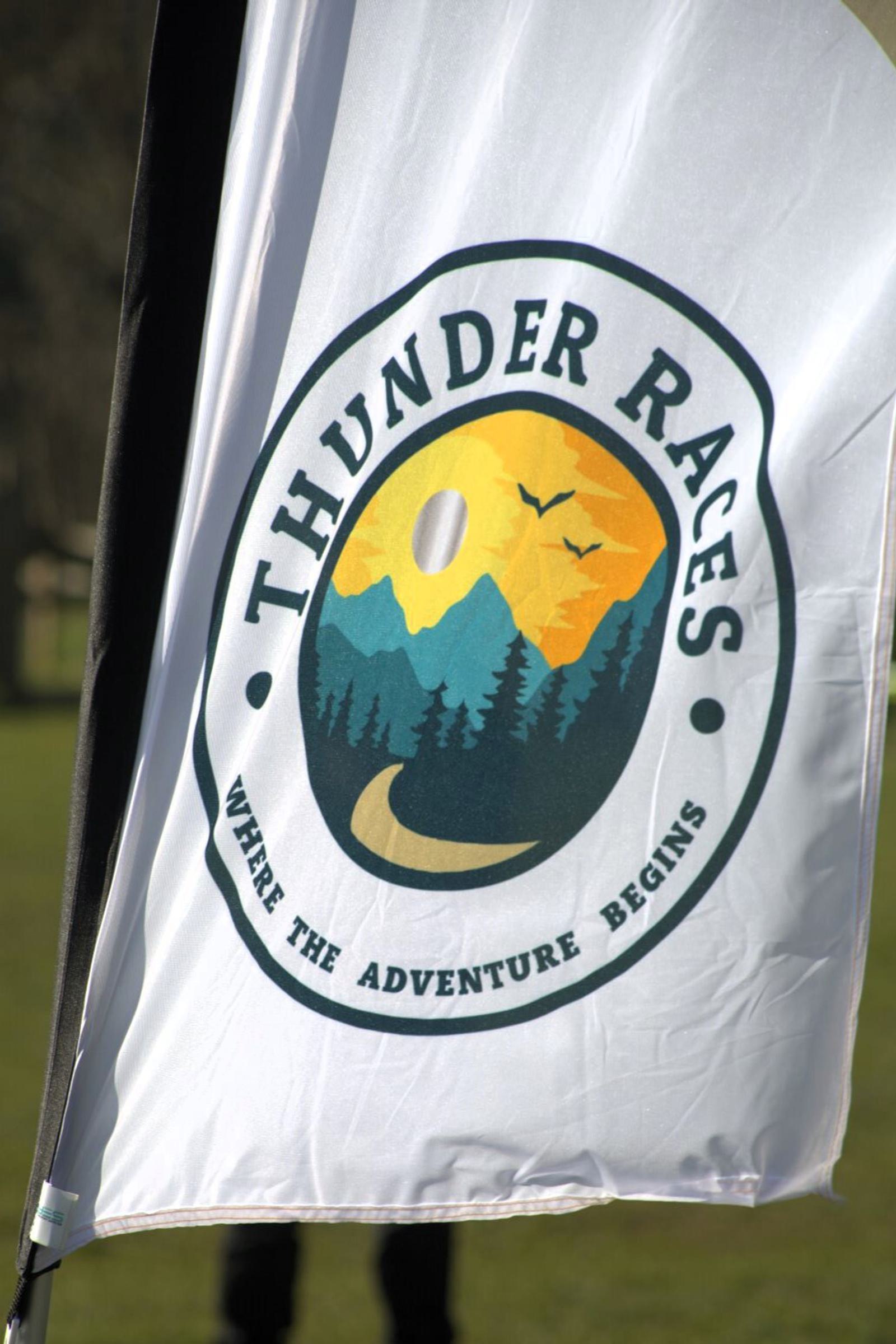 A white flag with the logo for "Thunder Races." The logo features a mountain, trees, and birds with the tagline "Where the Adventure Begins." The flag is outdoors, with green grass and a blurred background.