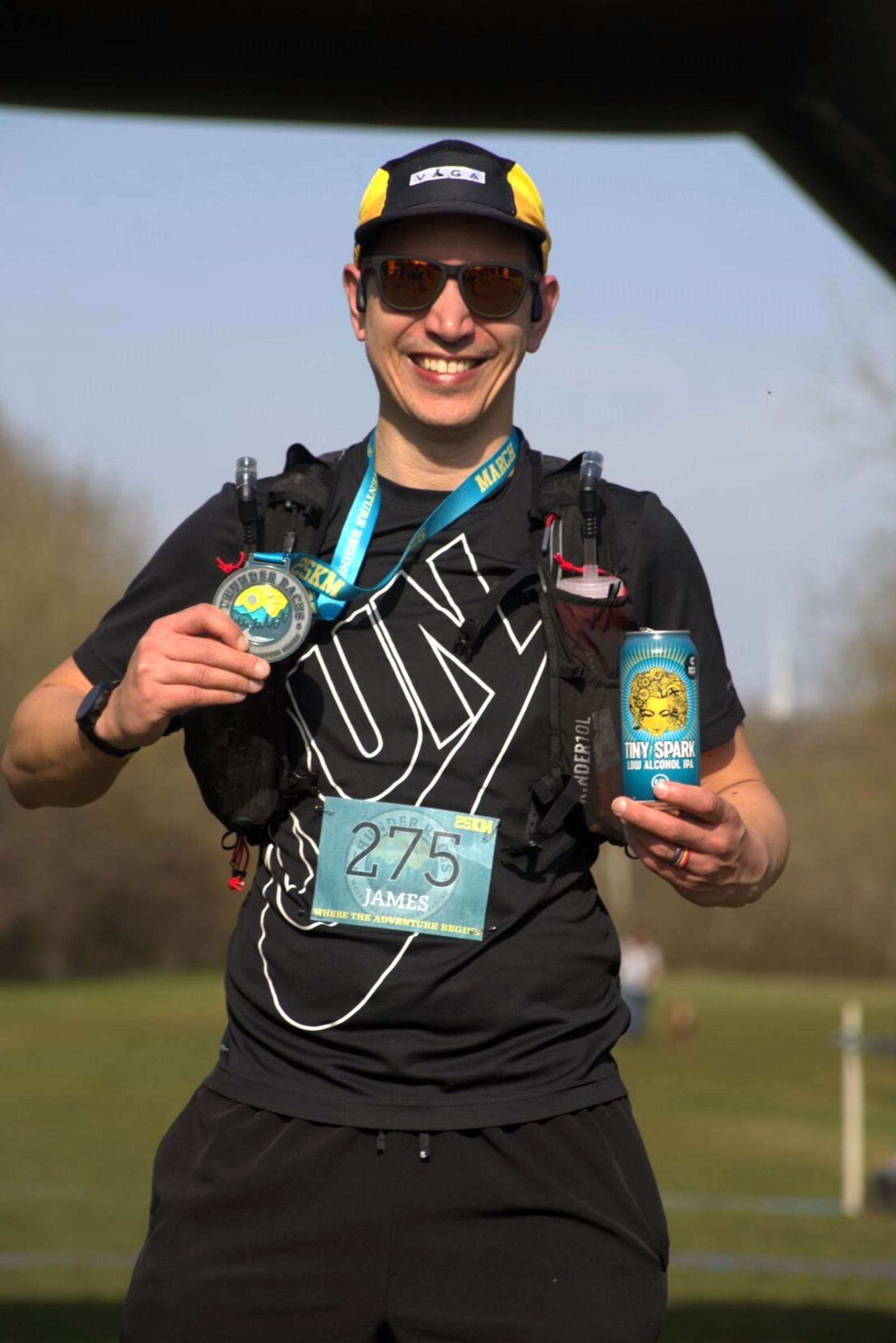 A person wearing a black athletic outfit and cap smiles and holds a medal and a can. They have a race number on their shirt and are outside on a sunny day with a green field in the background.