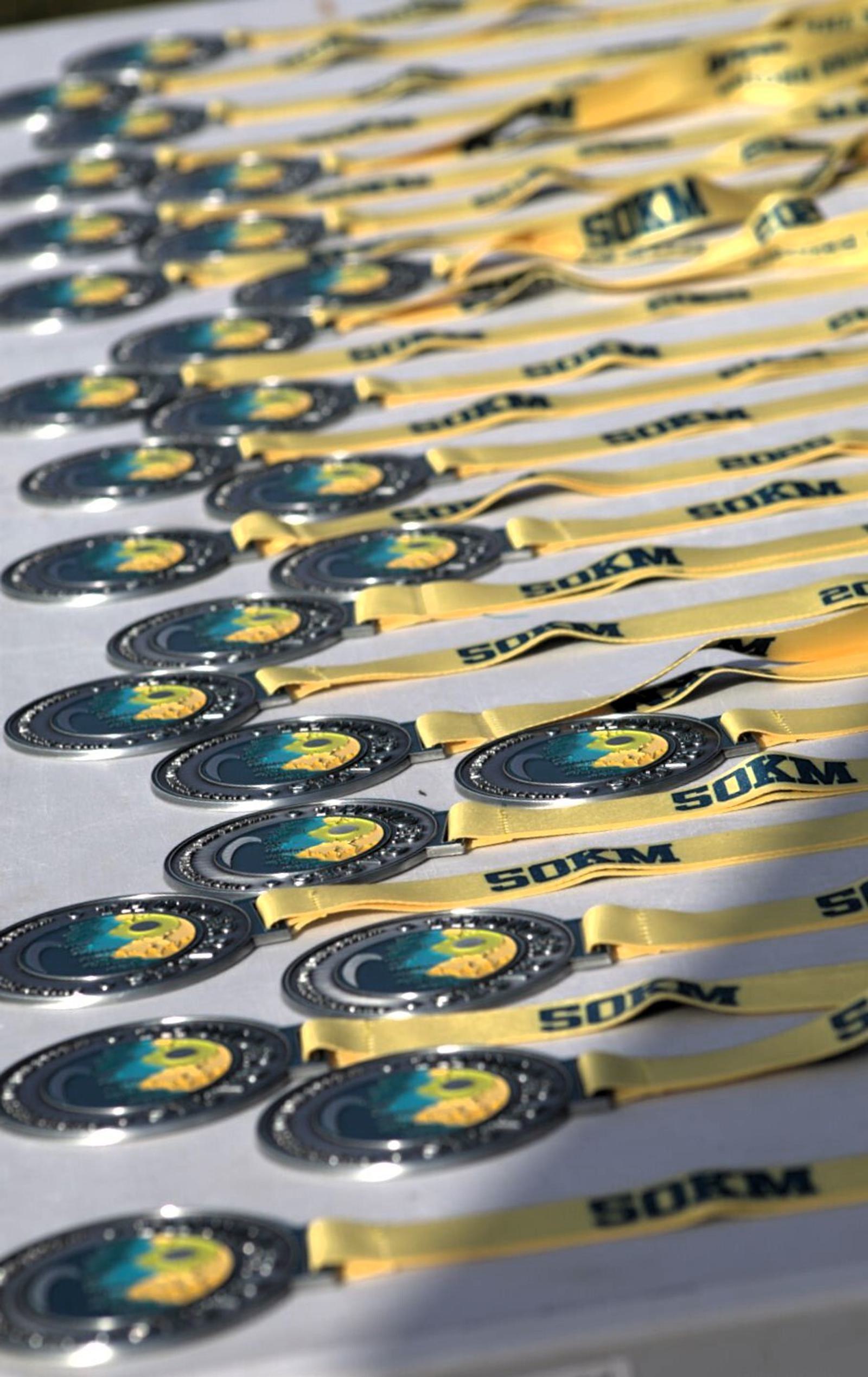 A table displays numerous silver medals with yellow ribbons. Each medal features a blue and green graphic and the text "SOKM" on the ribbon. They are arranged in straight rows under bright lighting.