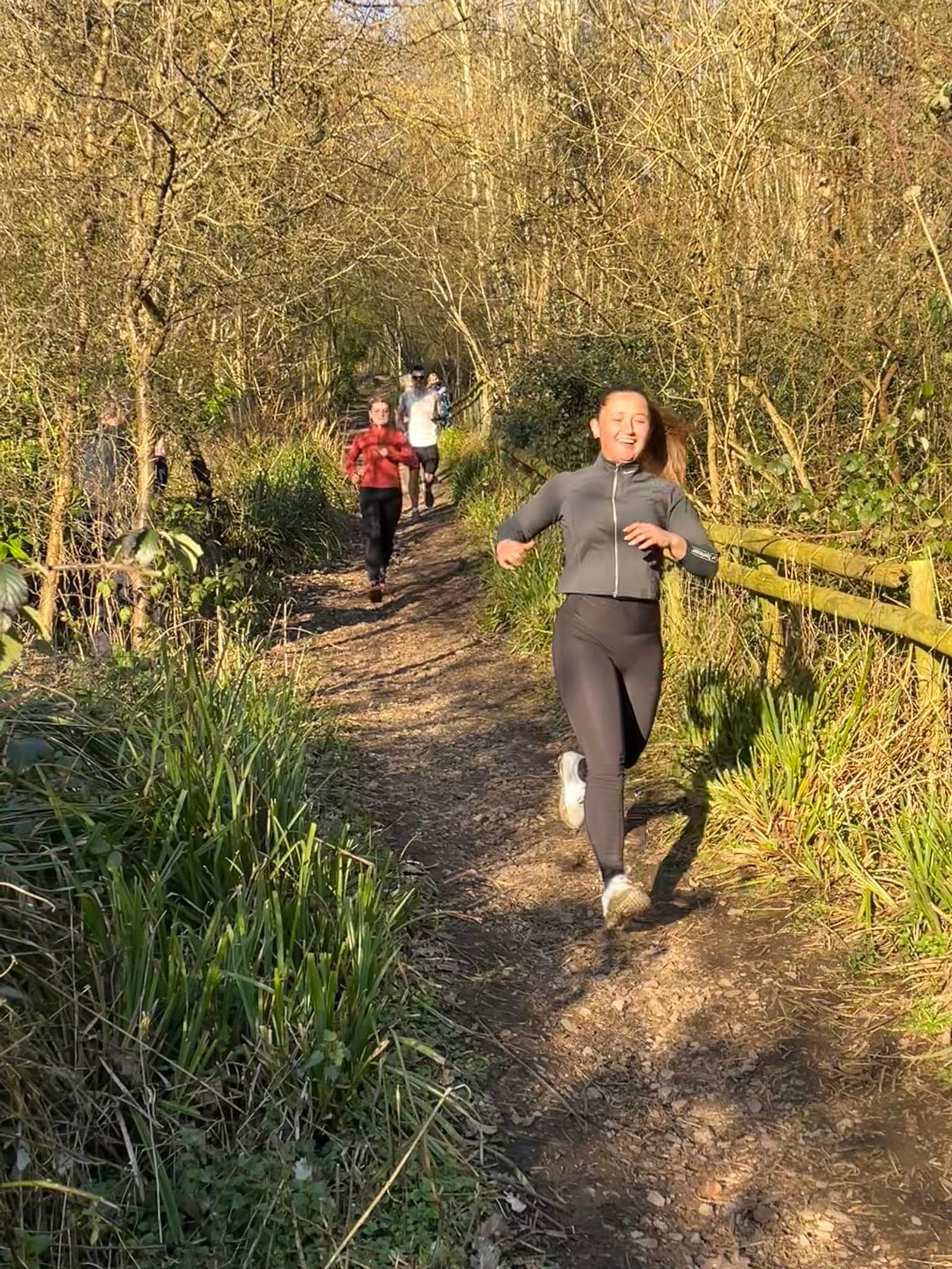 Three people are jogging on a woodland trail in bright sunlight. The person in front is wearing black activewear, followed by two others in lighter clothing. Trees and greenery surround the dirt path.