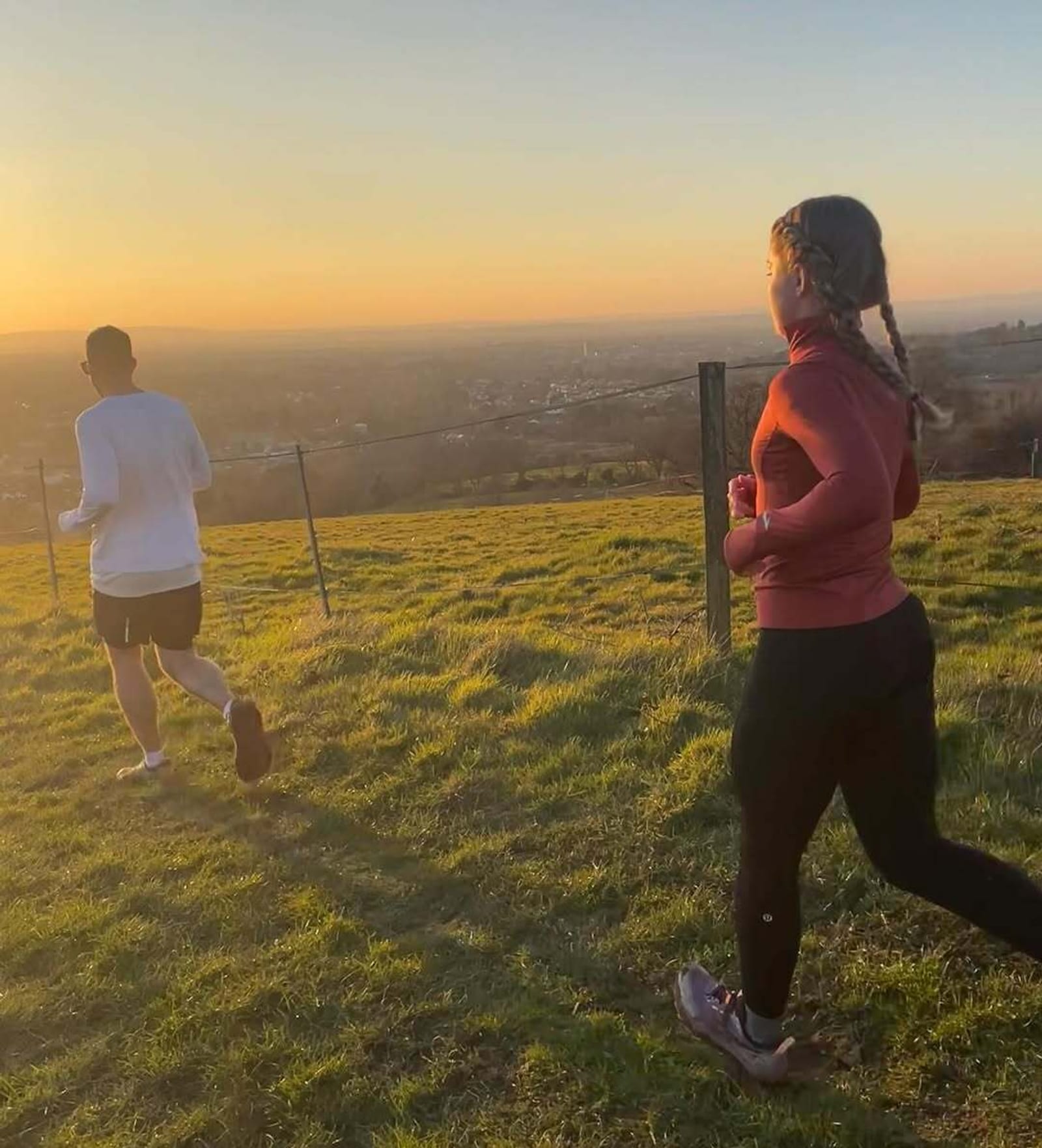 Two people run on a grassy hill at sunset. The person in front wears a white shirt and black shorts, while the person behind wears a red top and black pants. The golden sunlight casts long shadows over the landscape.