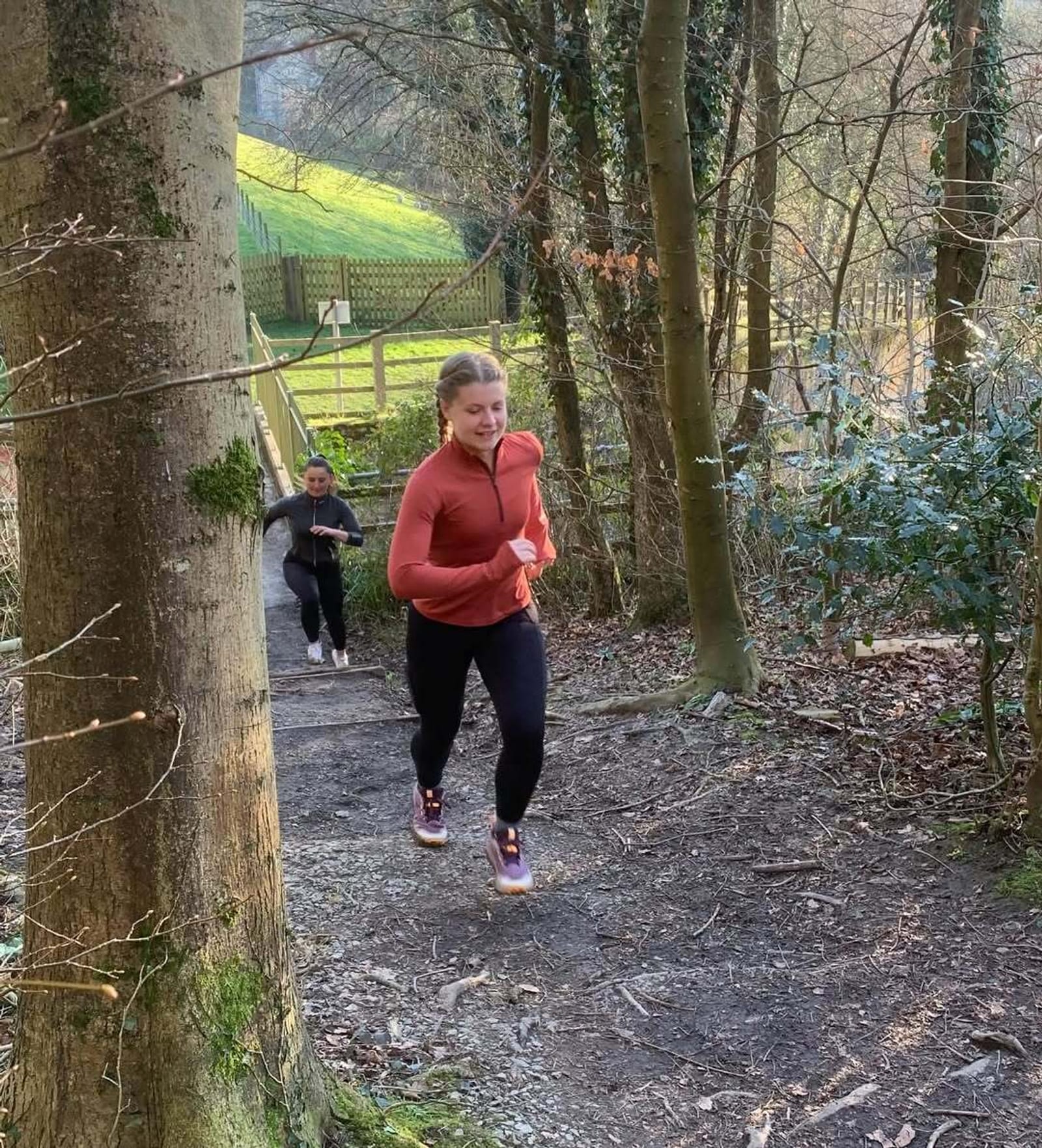 Two women are jogging up a dirt trail through a wooded area. One is in front wearing a red jacket, black leggings, and sneakers, while the other follows behind. Sunlight filters through the trees.