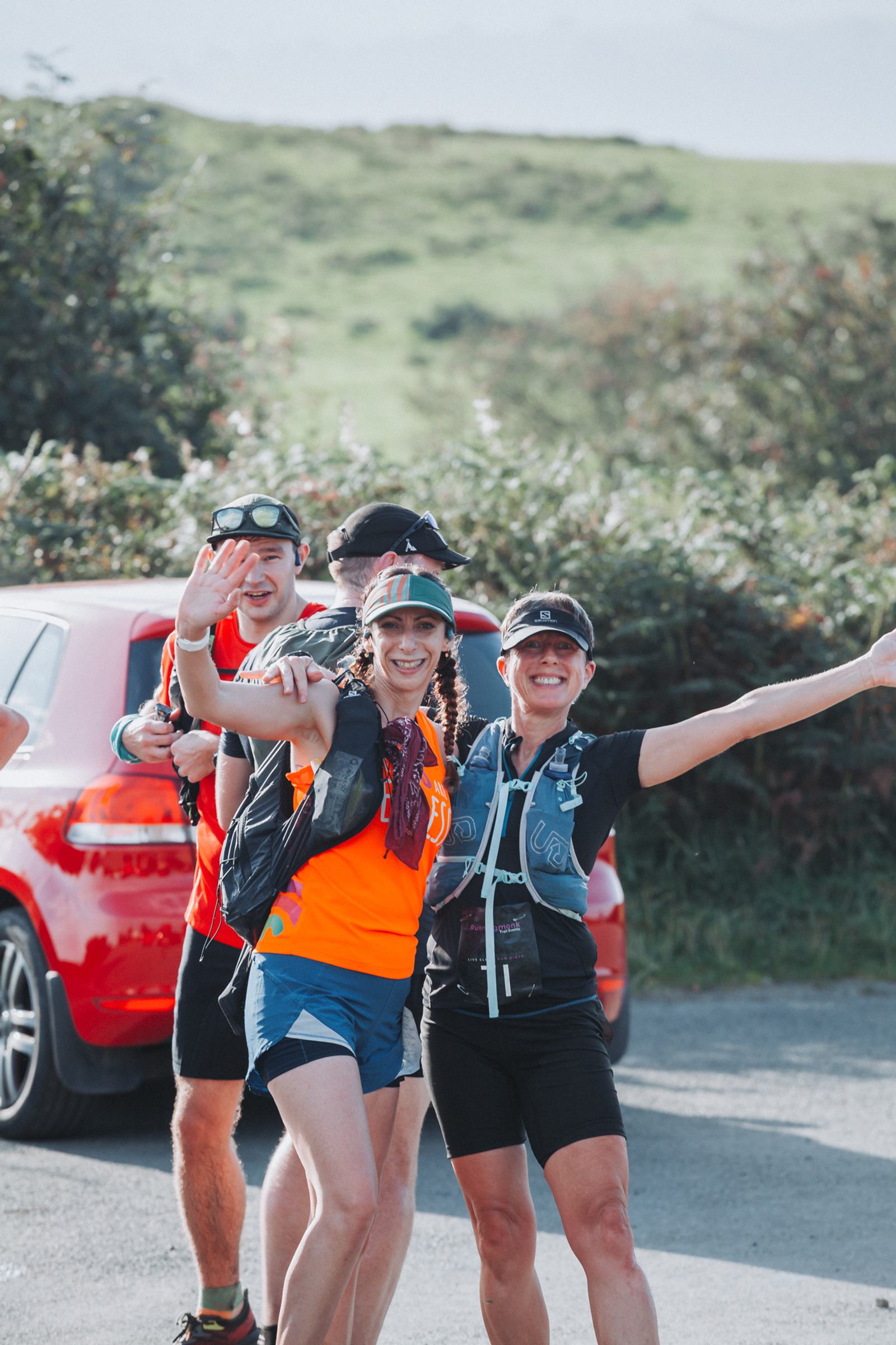 A group of runners in athletic gear, smiling and posing with arms outstretched near a red car. They are outdoors with greenery and a hill in the background.