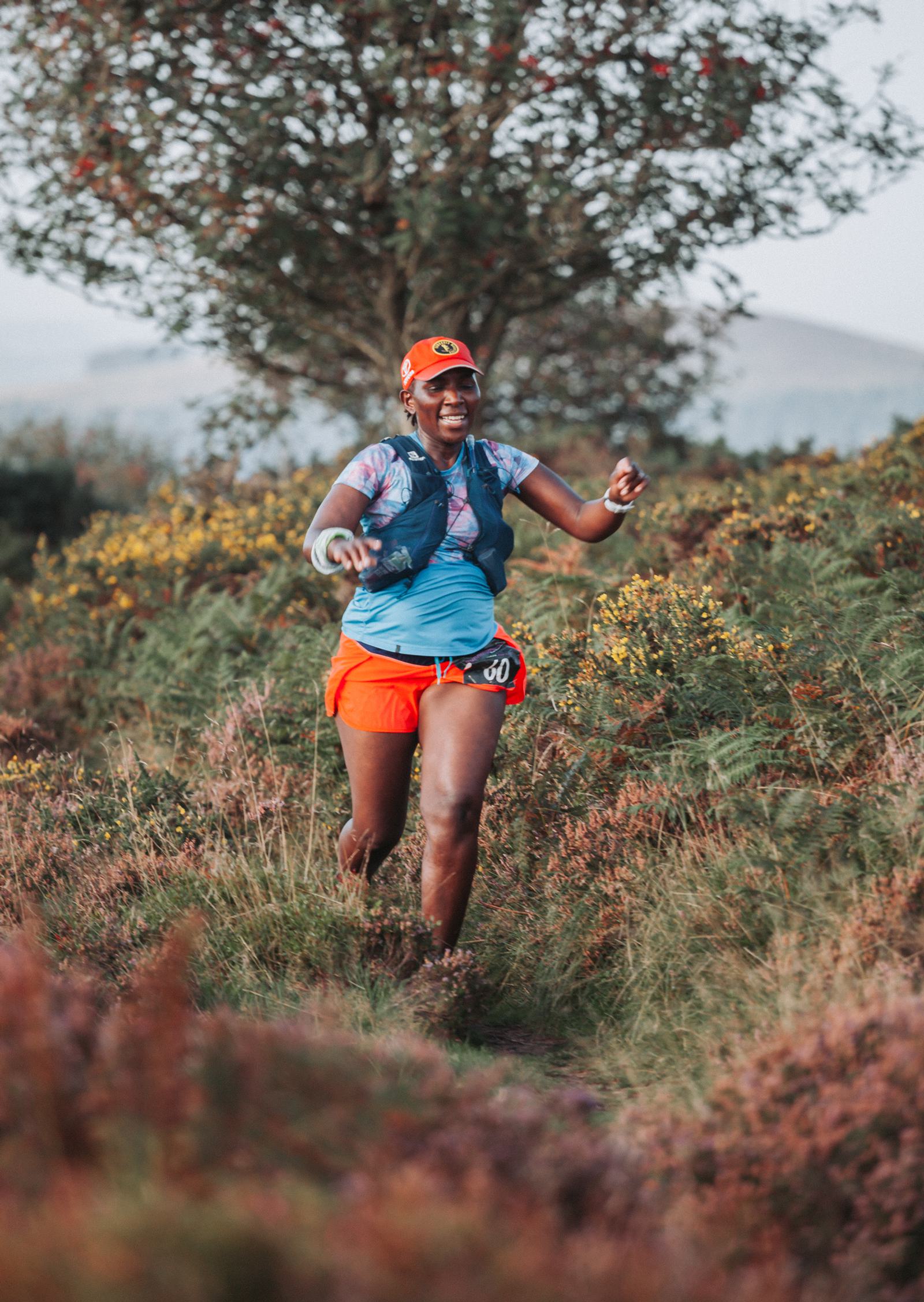 A runner wearing a red cap, a blue shirt, and orange shorts is smiling while jogging through a trail surrounded by tall grass and shrubs. A tree stands in the background against a hilly landscape.