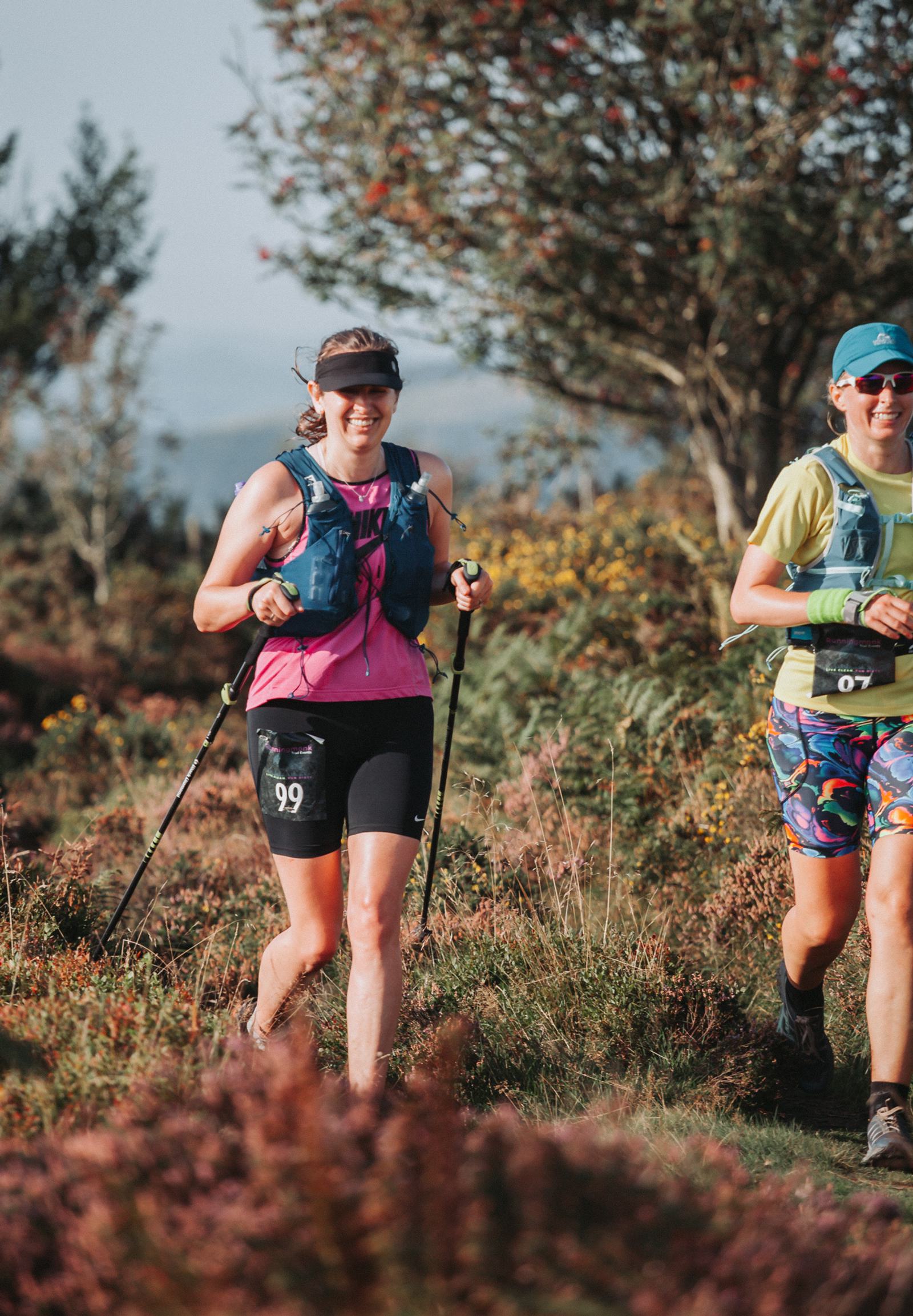 Two people are trail running on a scenic path surrounded by greenery and blooming heather. They wear athletic gear and backpacks, with one person using trekking poles. The sky is clear, suggesting a pleasant day.