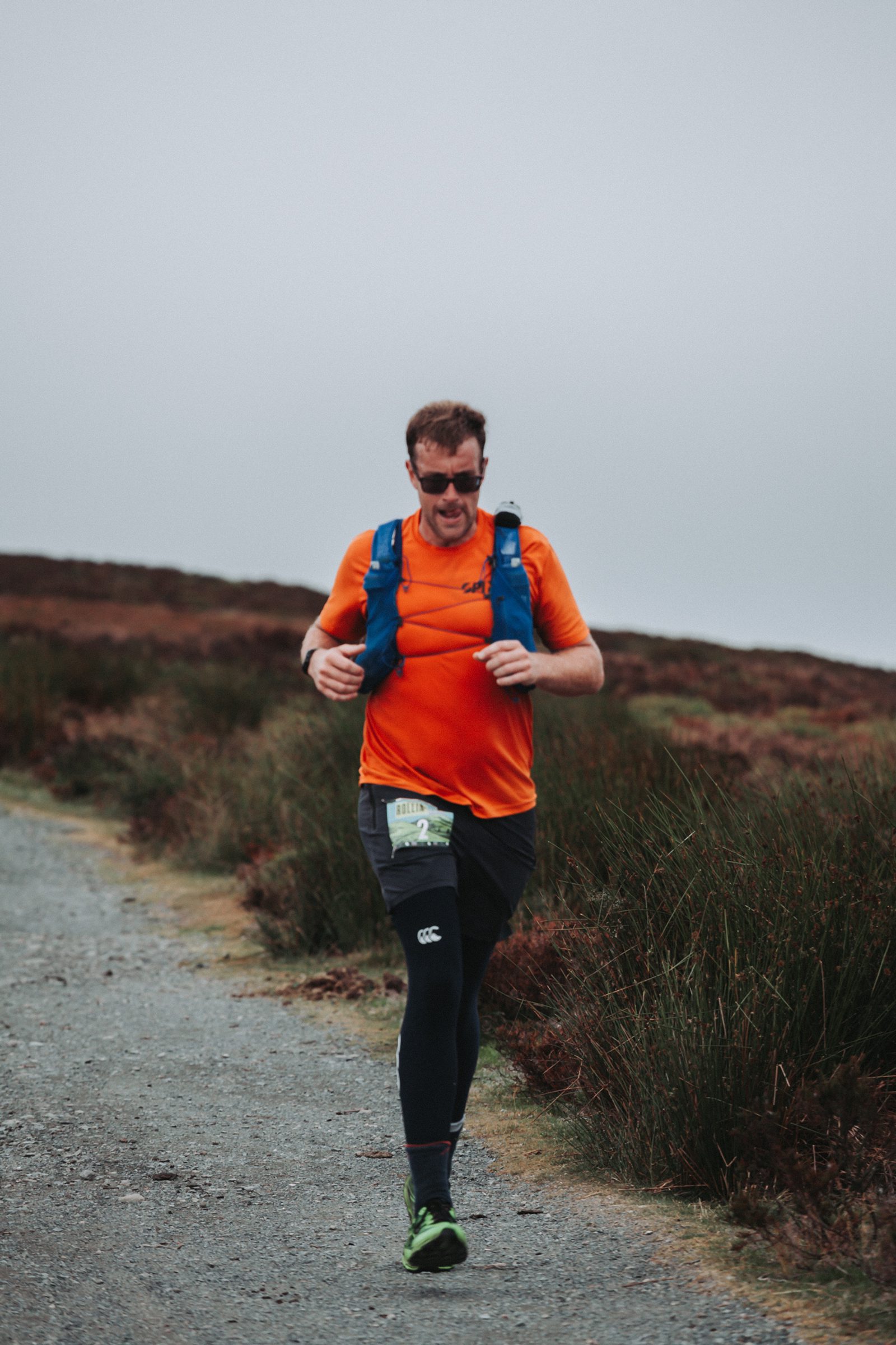 A man wearing an orange shirt, black leggings, and a blue running vest jogs on a gravel path through a grassy, hilly landscape under a cloudy sky. He has sunglasses and a race bib on his leg.