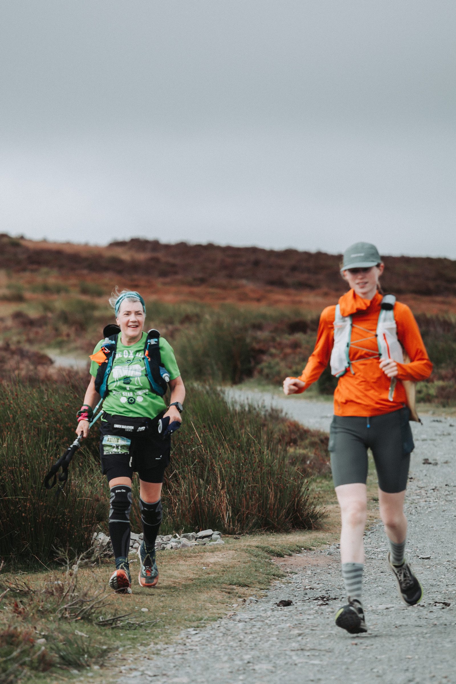Two people are hiking on a gravel path in a hilly outdoor setting. One wears a green shirt and black gear, smiling at the camera, while the other in orange and gray walks ahead, looking down. The sky is overcast.