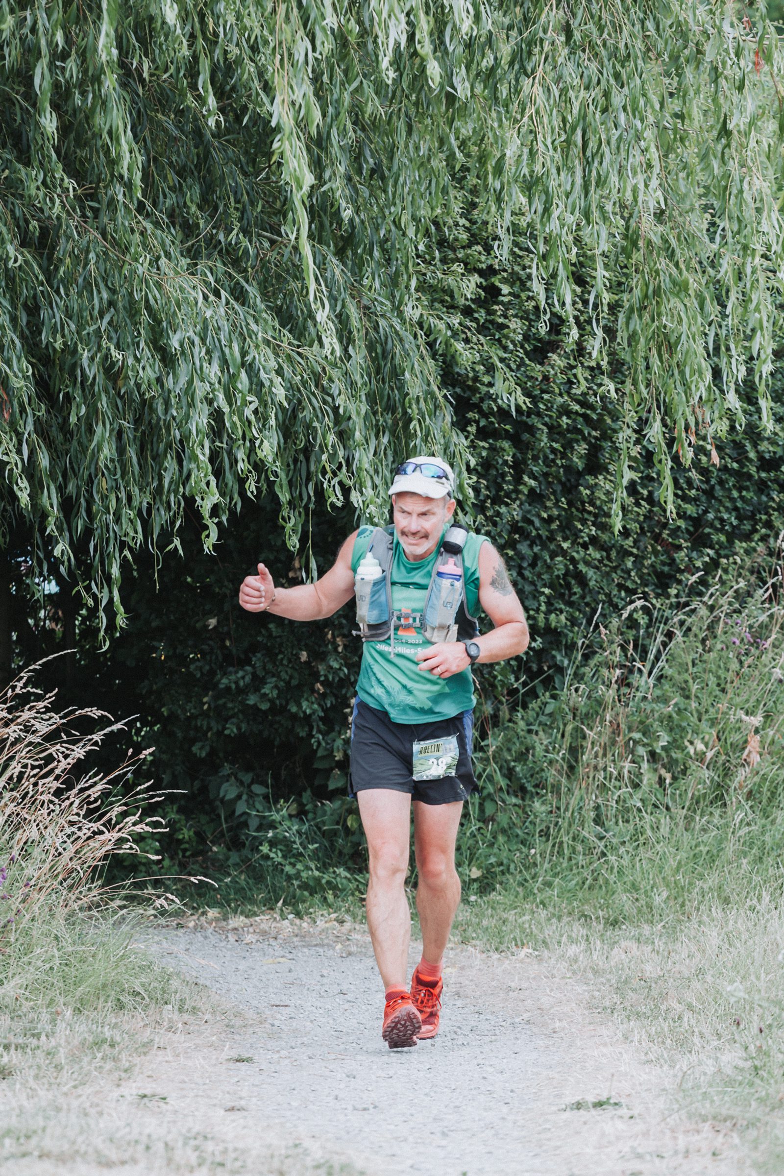 A runner wearing a green shirt, black shorts, and a hydration vest smiles and gives a thumbs up while jogging on a trail surrounded by greenery and tall grass.