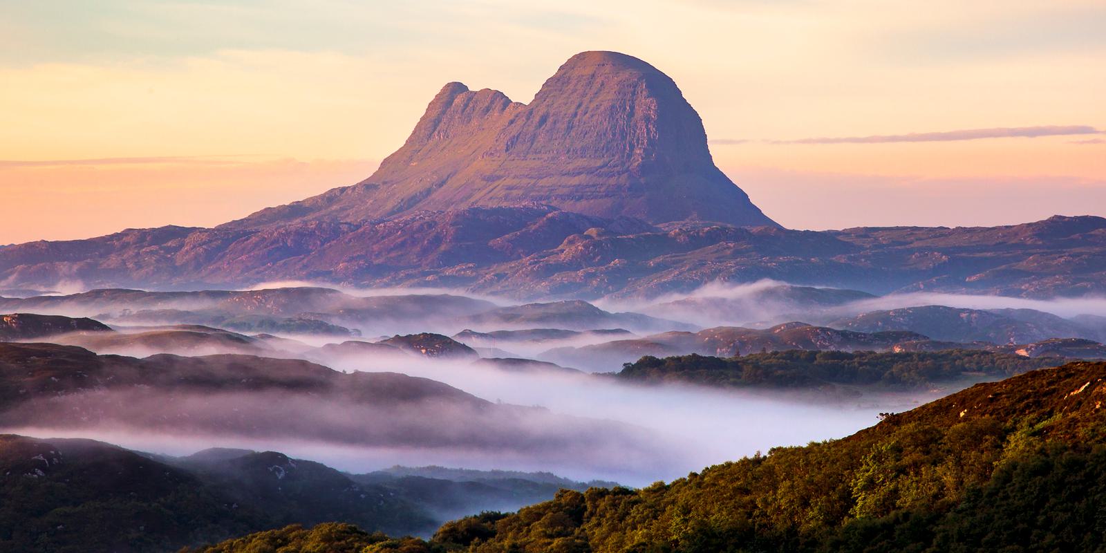 A stunning view of Suilven mountain in Scotland at sunrise. The peak rises majestically above a landscape of rolling hills and valleys, partially shrouded in mist. The sky is a soft blend of pink and orange hues.