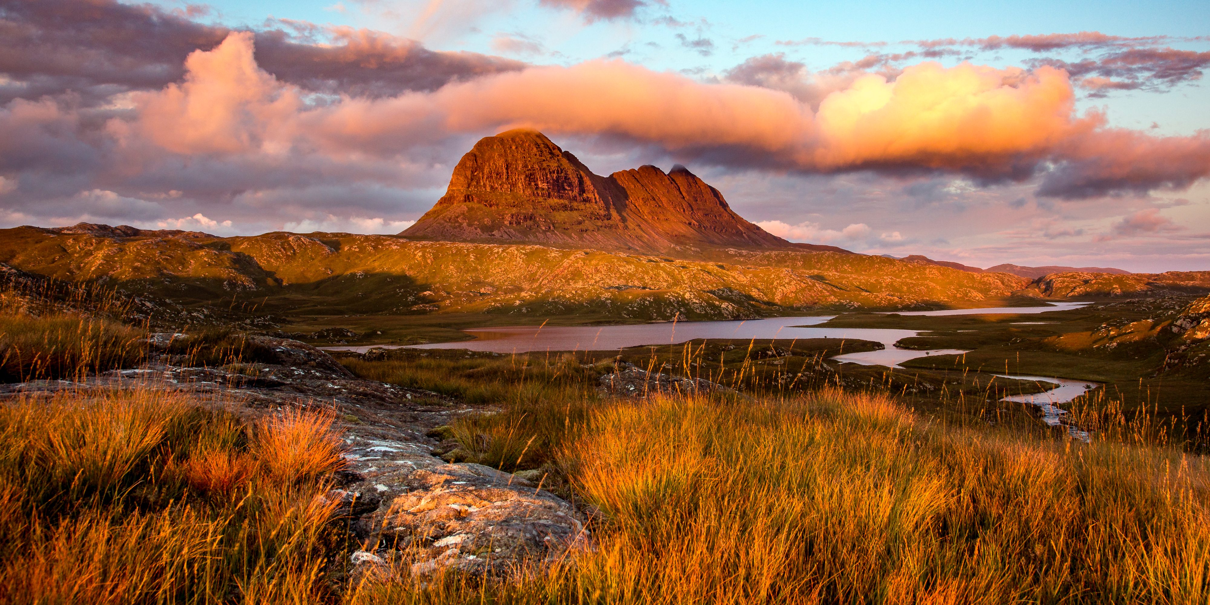 A stunning sunset casts a warm glow over a mountain landscape with grassy foreground. The sky is partly cloudy, reflecting orange and pink hues, and a winding river or lake nestles at the mountain's base.