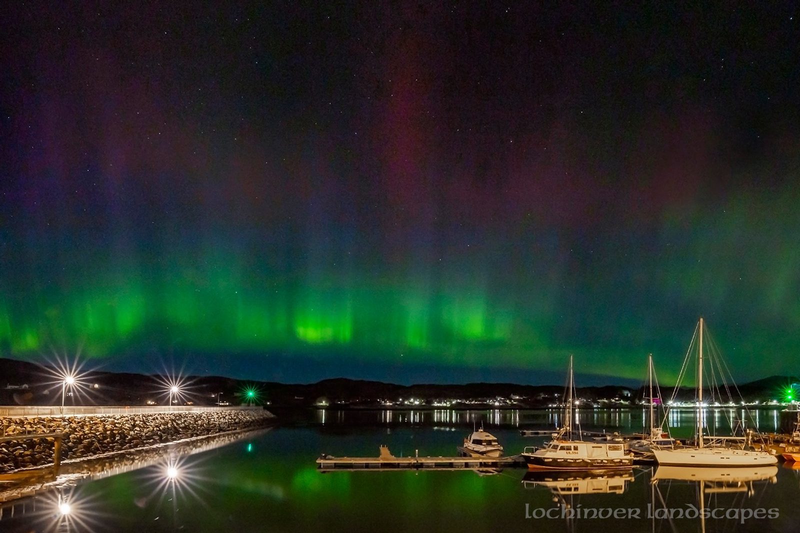 The northern lights glow green and purple in the night sky above a calm marina with docked boats, their reflections shimmering in the water. The scene is peaceful and illuminated by harbor lights.