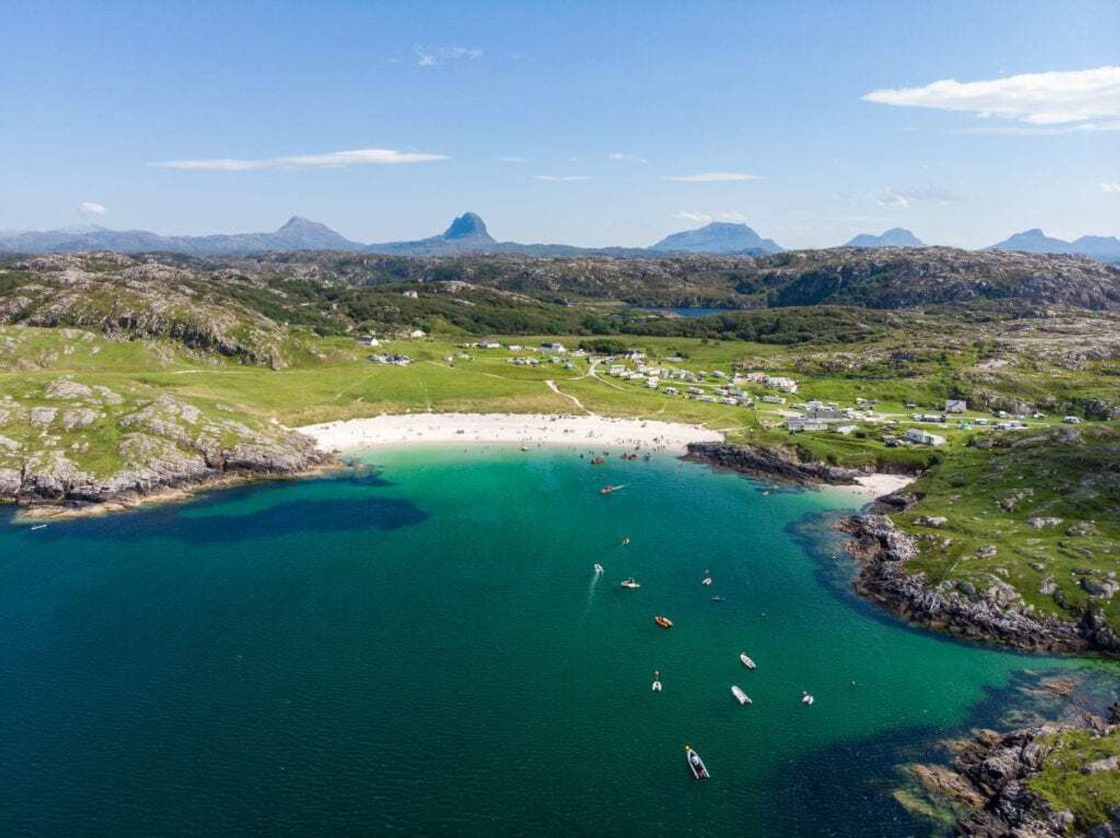 Aerial view of a scenic bay with turquoise water, a sandy beach, scattered boats, and a small village surrounded by green hills and distant mountains under a blue sky.