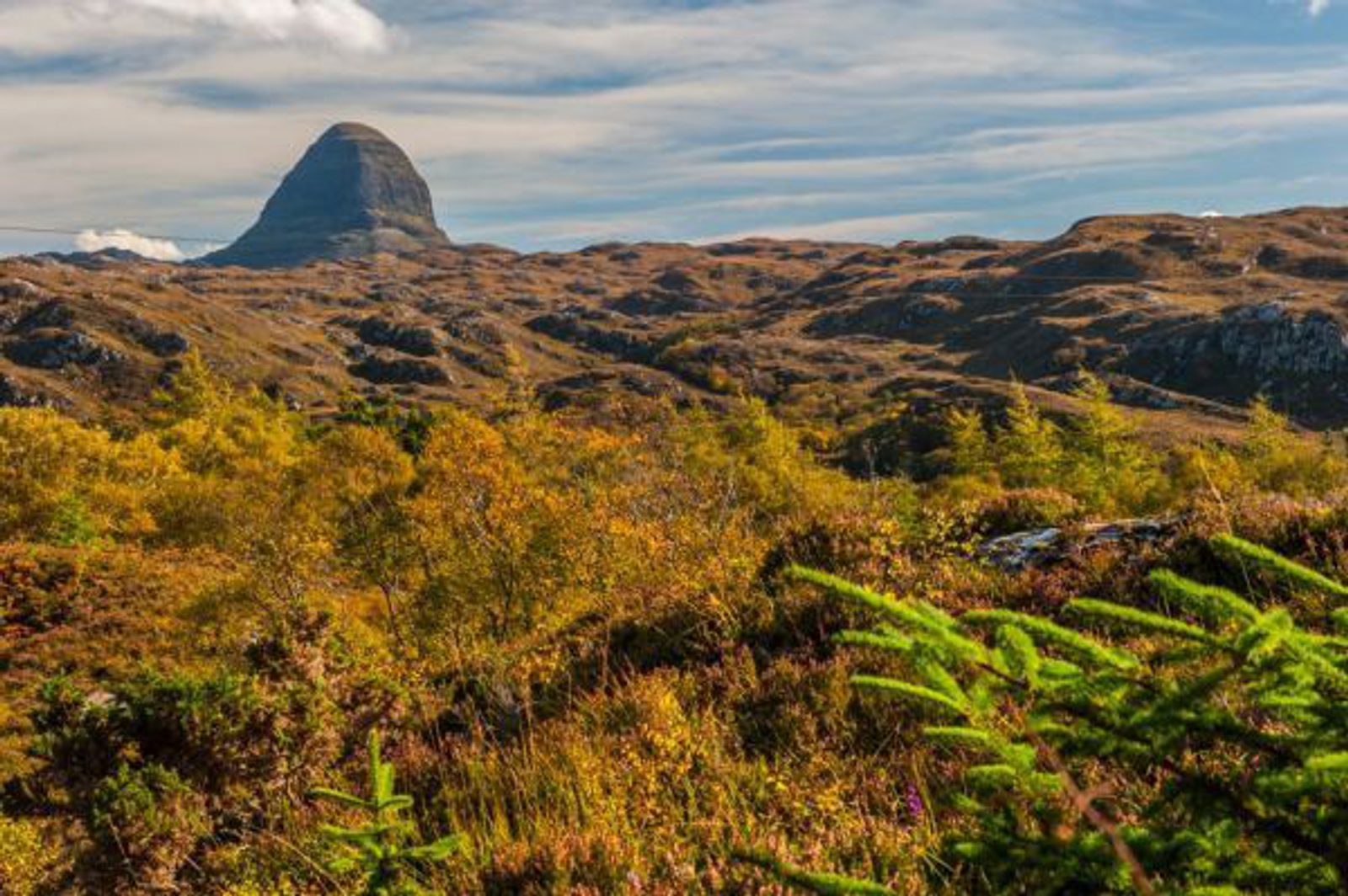 A rugged, rocky mountain rises in the background under a partly cloudy sky, with a foreground of green and golden autumn foliage and rolling hills.