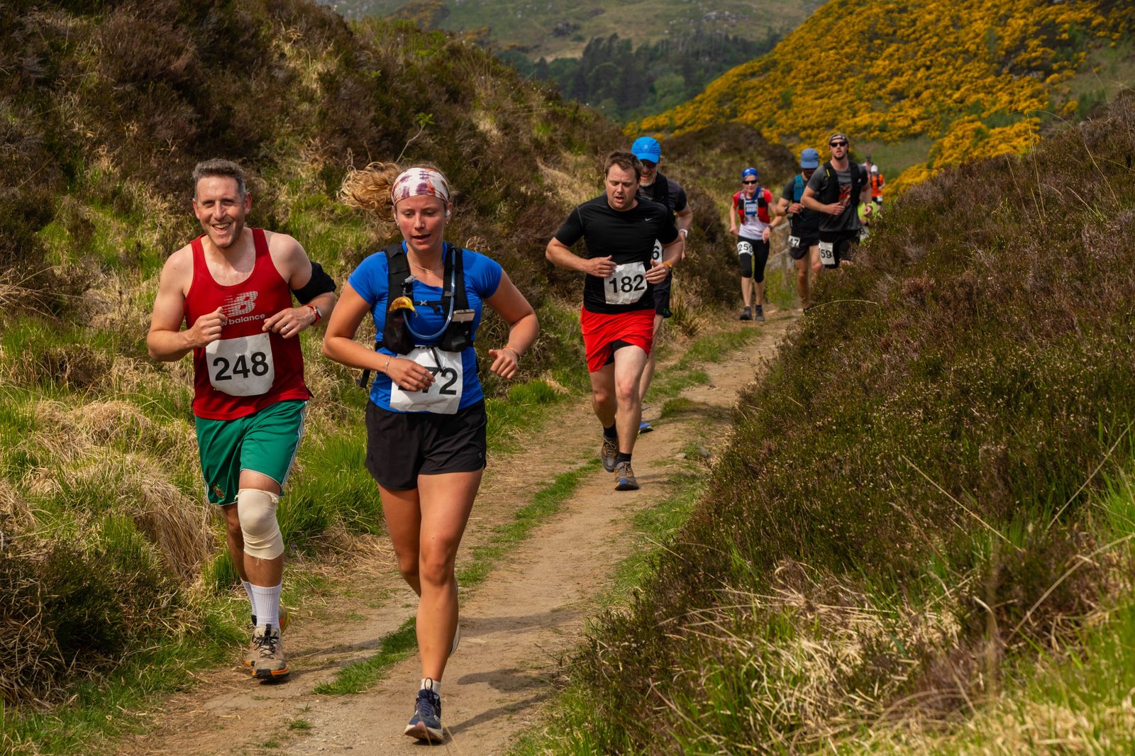 Runners with race numbers jog along a narrow trail through grassy, hilly terrain on a sunny day, with bushes and yellow flowers in the background. The two in front are smiling and wearing athletic gear.