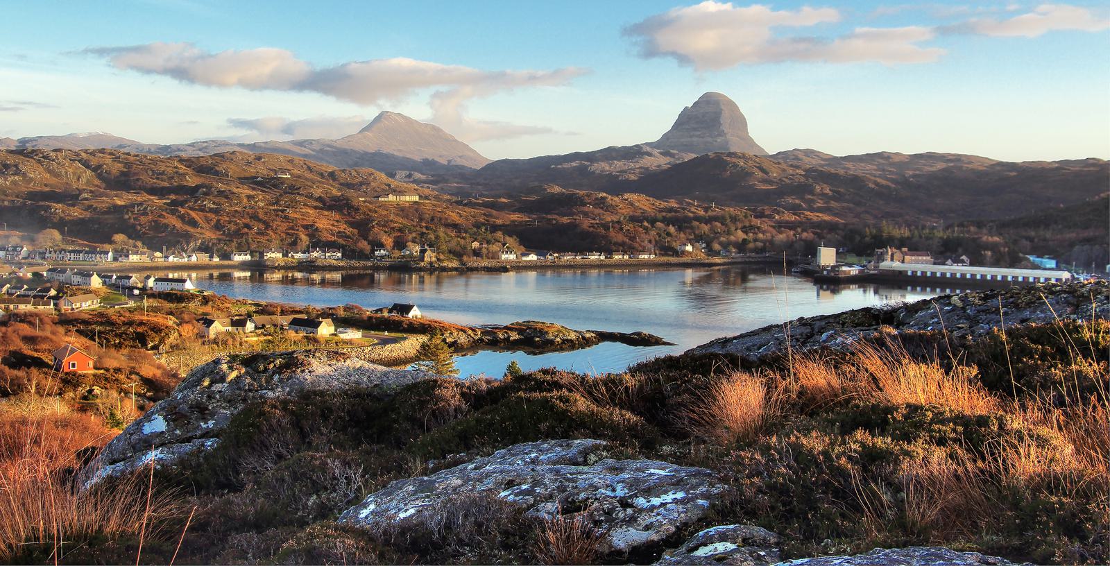 A serene landscape featuring a coastal village with small houses near a calm body of water. Rolling hills and dramatic mountains rise in the background. The scene is bathed in warm, golden sunlight under a partly cloudy sky.