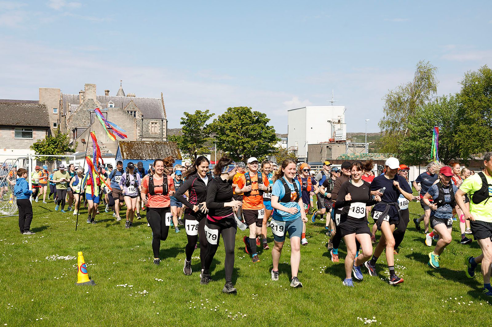 A group of runners in numbered bibs start a race on a grassy field under a sunny sky, with colorful flags, spectators, and buildings in the background.
