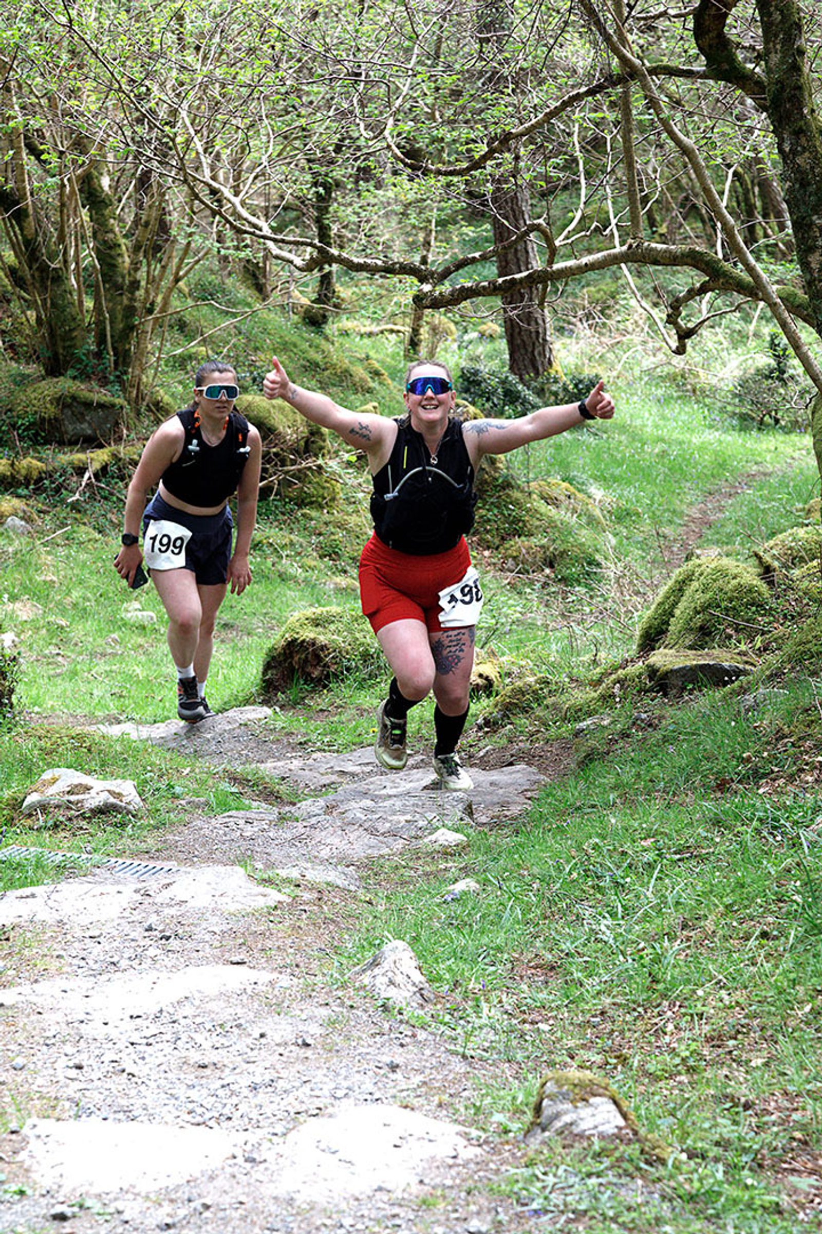Two trail runners, both wearing race bibs and sunglasses, are running uphill on a rocky forest path. The runner in front is smiling and giving two thumbs up, while the runner behind is smiling and following closely.