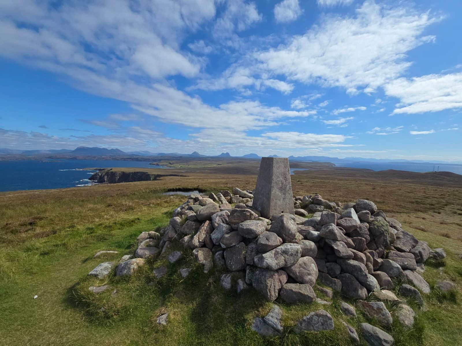 A stone cairn with a concrete trig point stands on grassy terrain overlooking cliffs, sea, and distant hills under a partly cloudy blue sky.