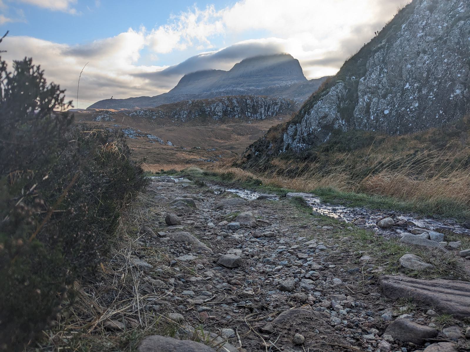 A rocky trail leads through a scenic landscape of grassy hills and large boulders under a partly cloudy sky. A mountain peak is visible in the background, partially shrouded in low-hanging clouds.