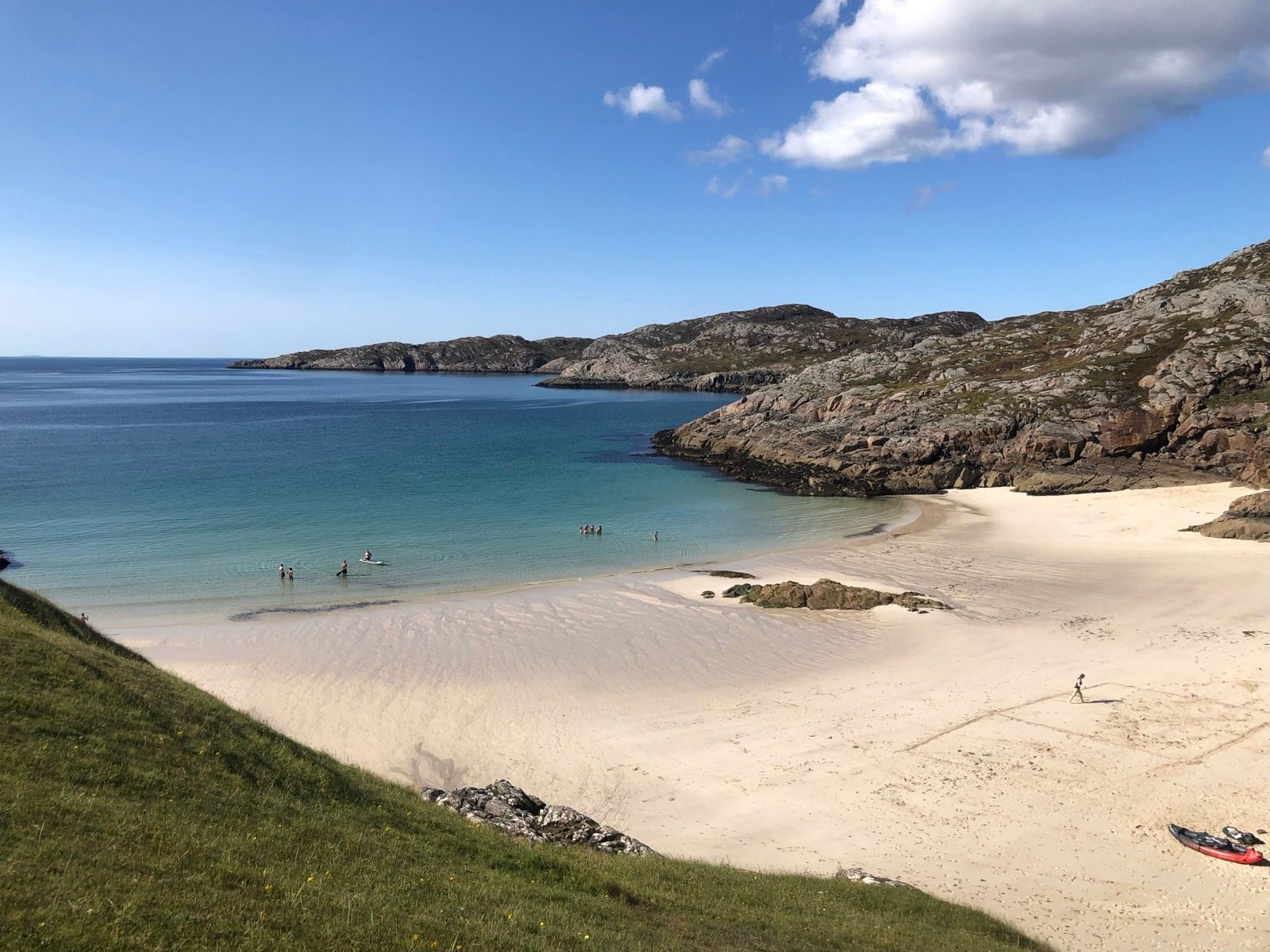 A sandy beach with clear blue water, a few people swimming, rocky hills in the background, and a person walking near a kayak on the shore under a bright, sunny sky.