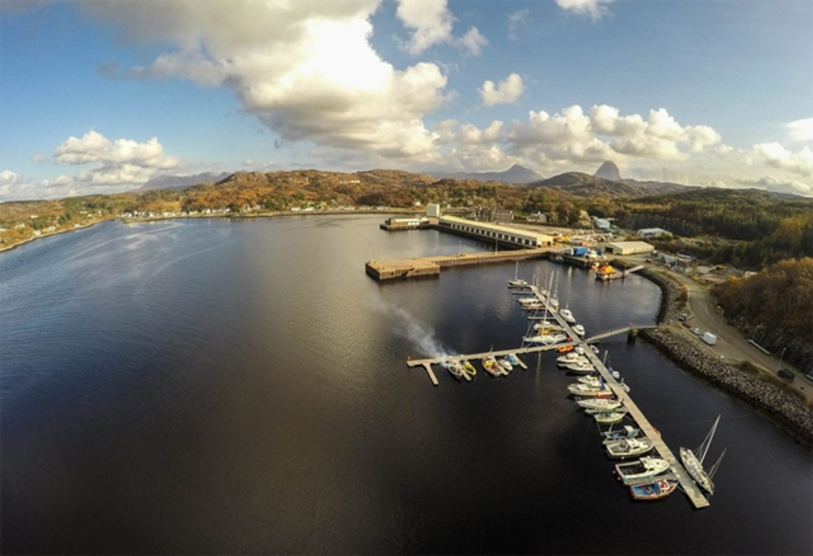 Aerial view of a marina with several boats docked along a pier, set on a calm bay surrounded by wooded hills and scattered buildings under a partly cloudy sky.