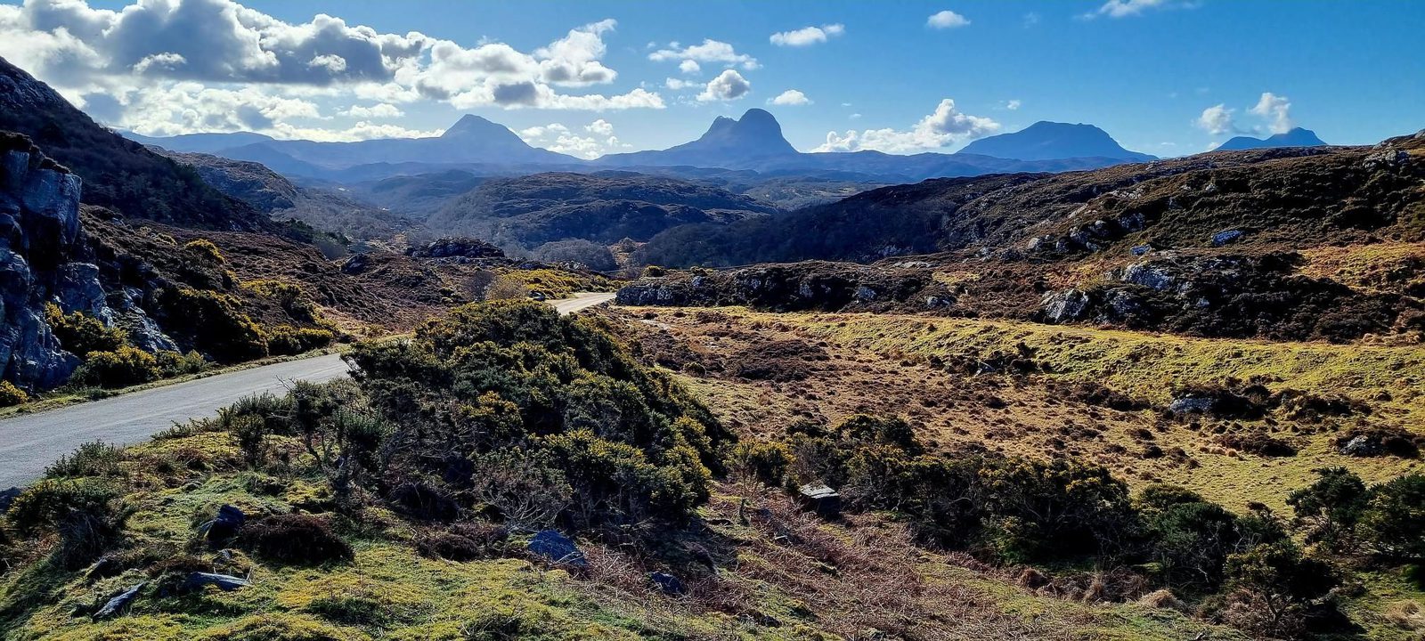 A winding road cuts through a rugged, grassy landscape with rolling hills and distant mountains under a blue sky dotted with fluffy clouds.