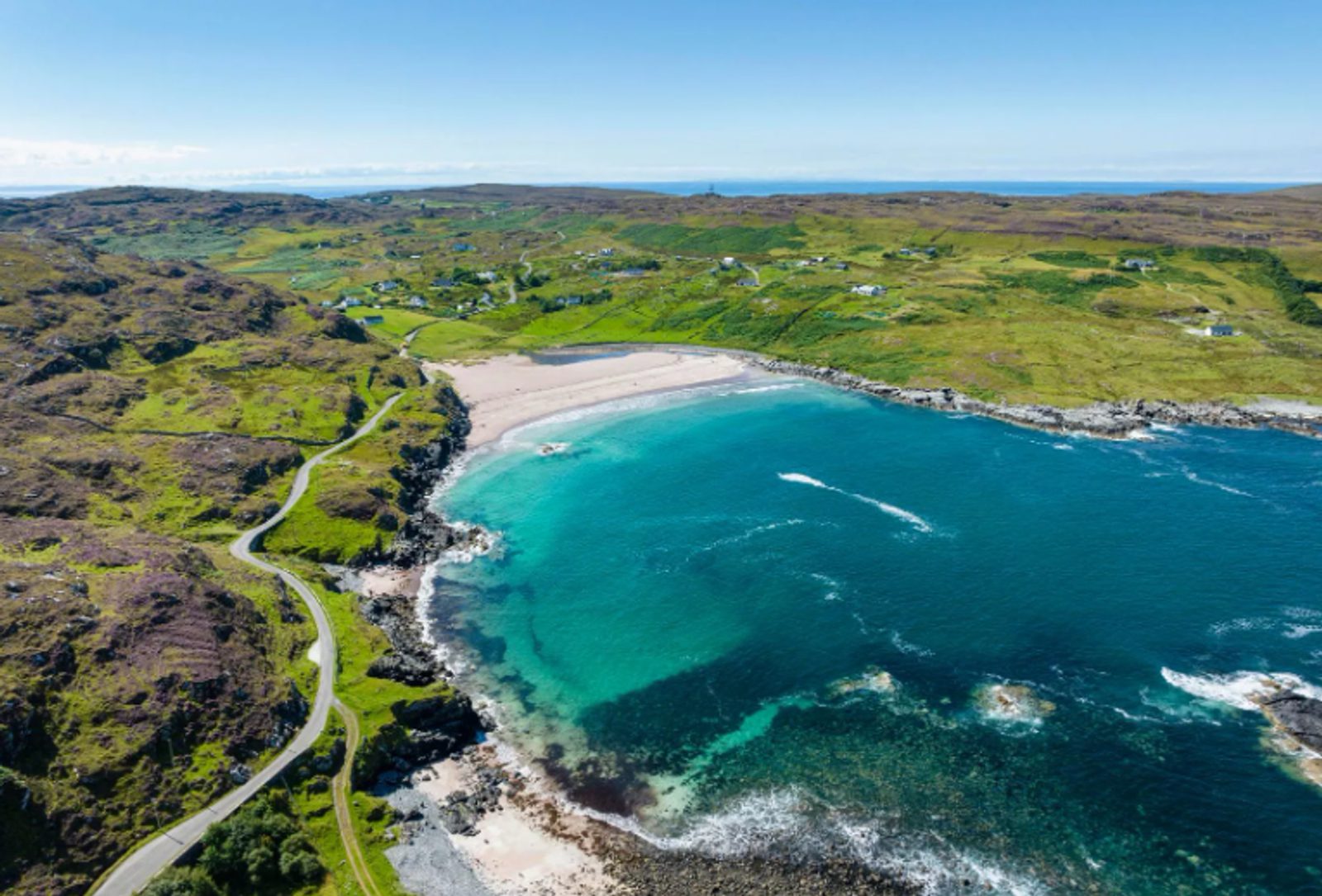 Aerial view of a curved sandy beach with turquoise water, surrounded by green hills, rocky shoreline, a winding coastal road, and scattered houses under a clear blue sky.