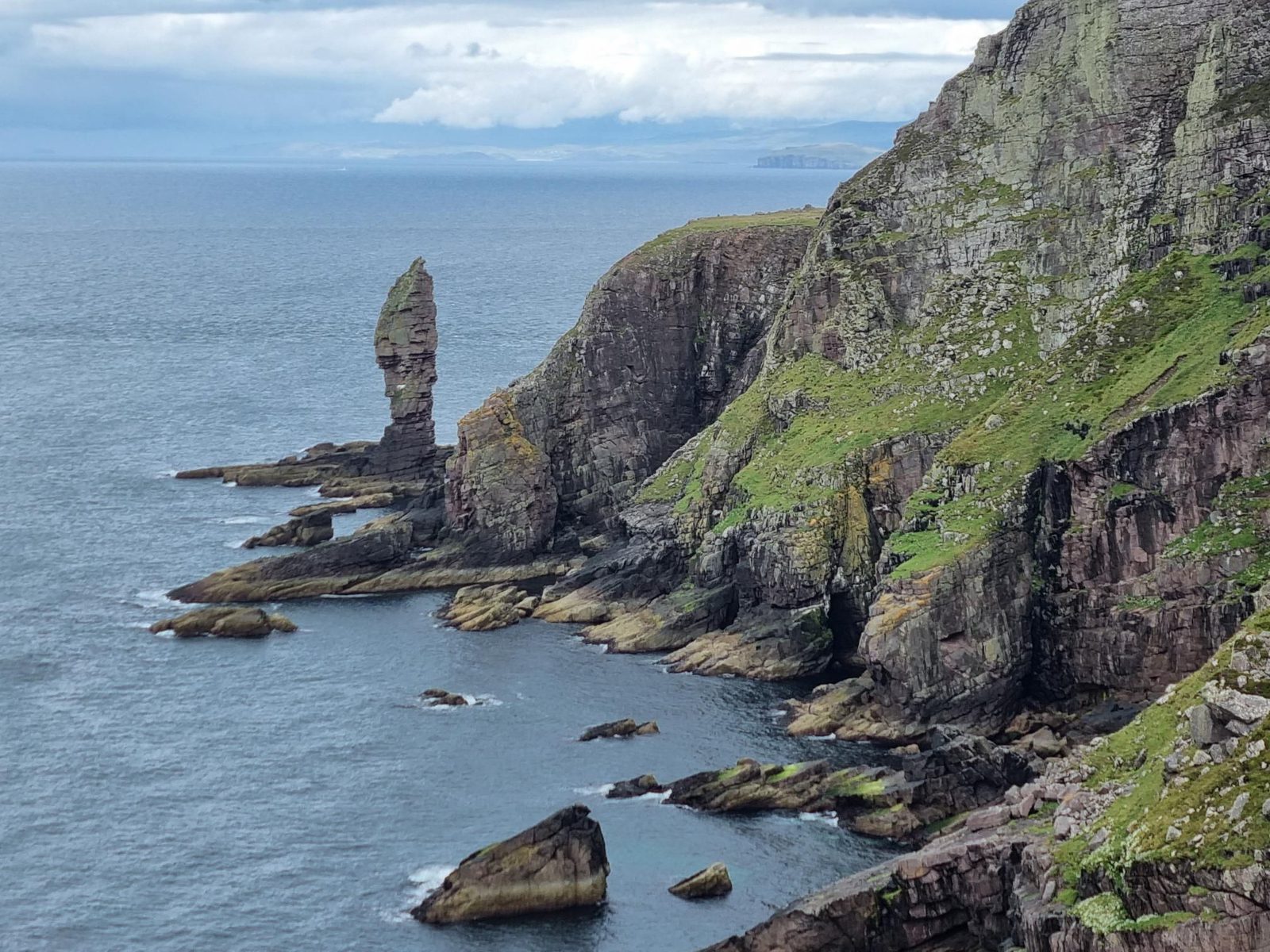 Rocky sea cliffs covered in green vegetation rise sharply from the water, with a tall, slender rock formation jutting up near the shoreline. The ocean stretches into the distance under a partly cloudy sky.