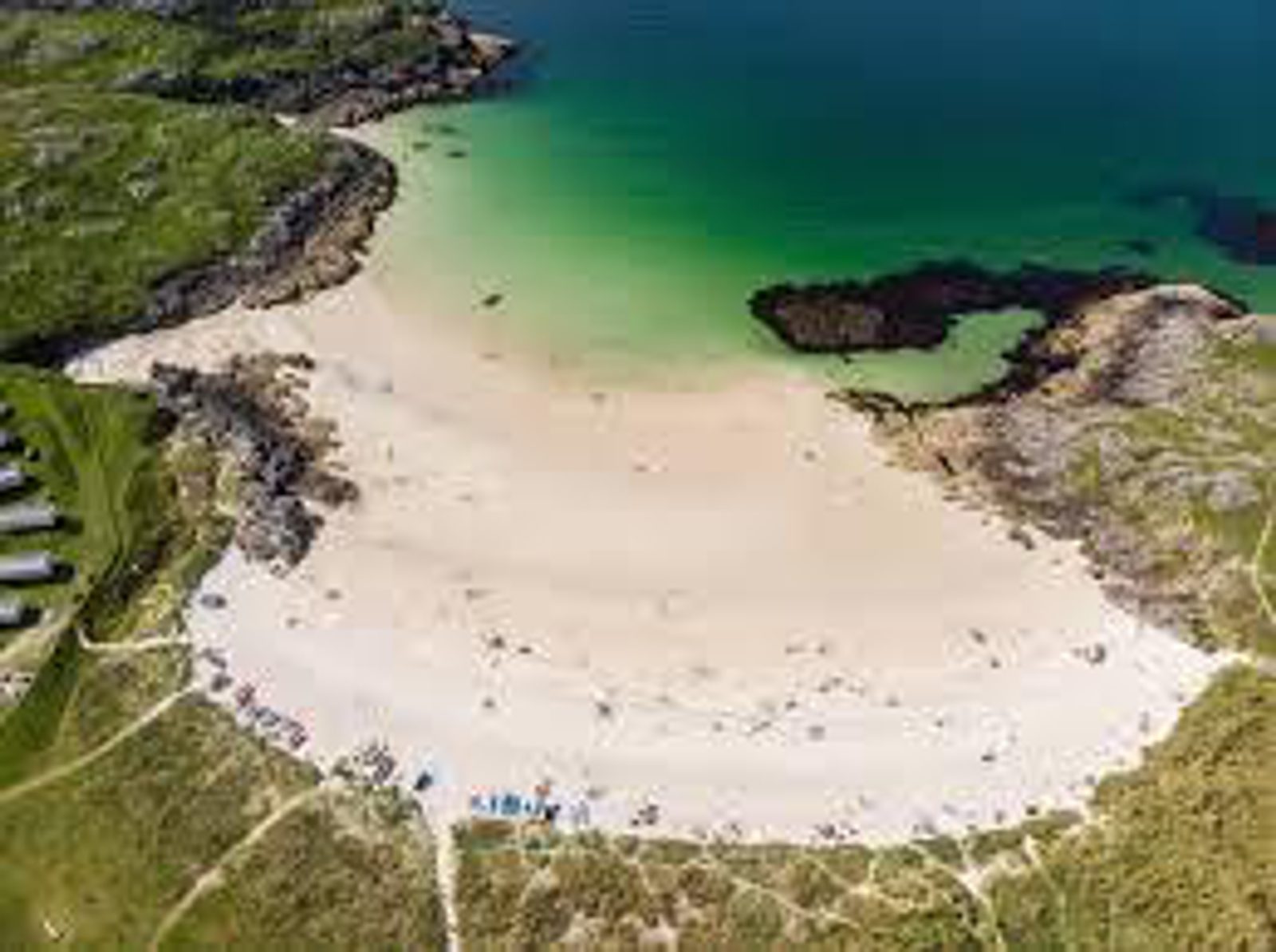 Aerial view of a sandy beach with turquoise water, bordered by green grass and rocky areas, with people and pathways visible along the shore.