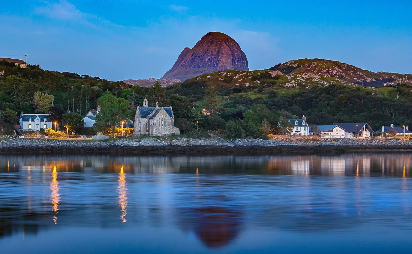 A calm body of water reflects lights from a small village with stone buildings and a church, with a prominent rounded mountain rising in the background under a clear blue sky.