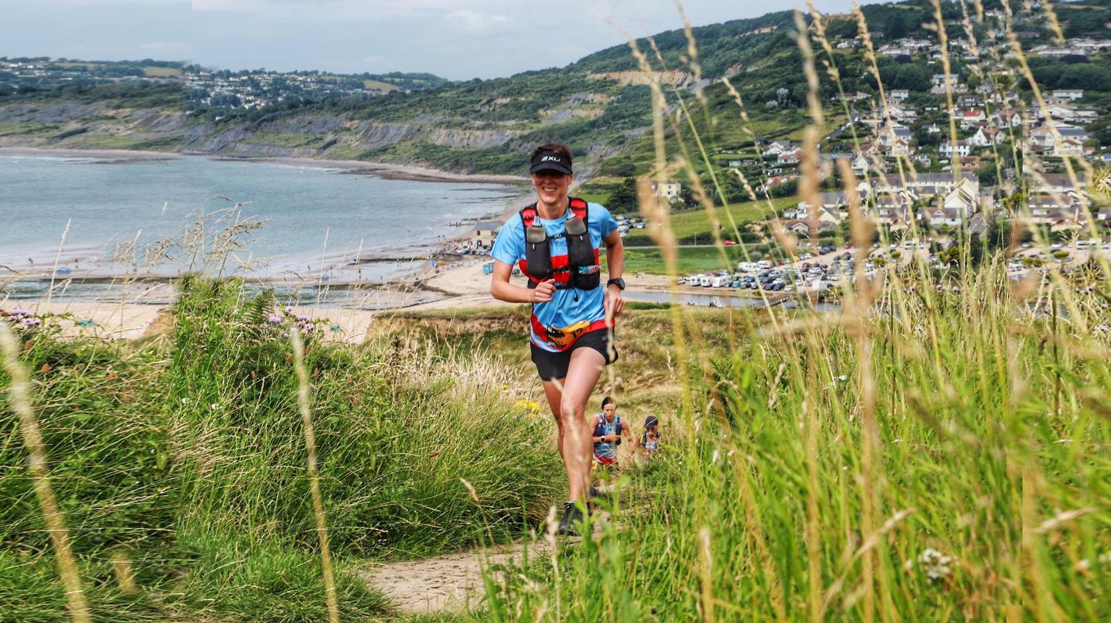 A person in a blue shirt and red backpack runs on a grassy coastal trail with a bay and village in the background. The sea is visible to the left, and hills and houses are on the right. Tall grass lines the path.
