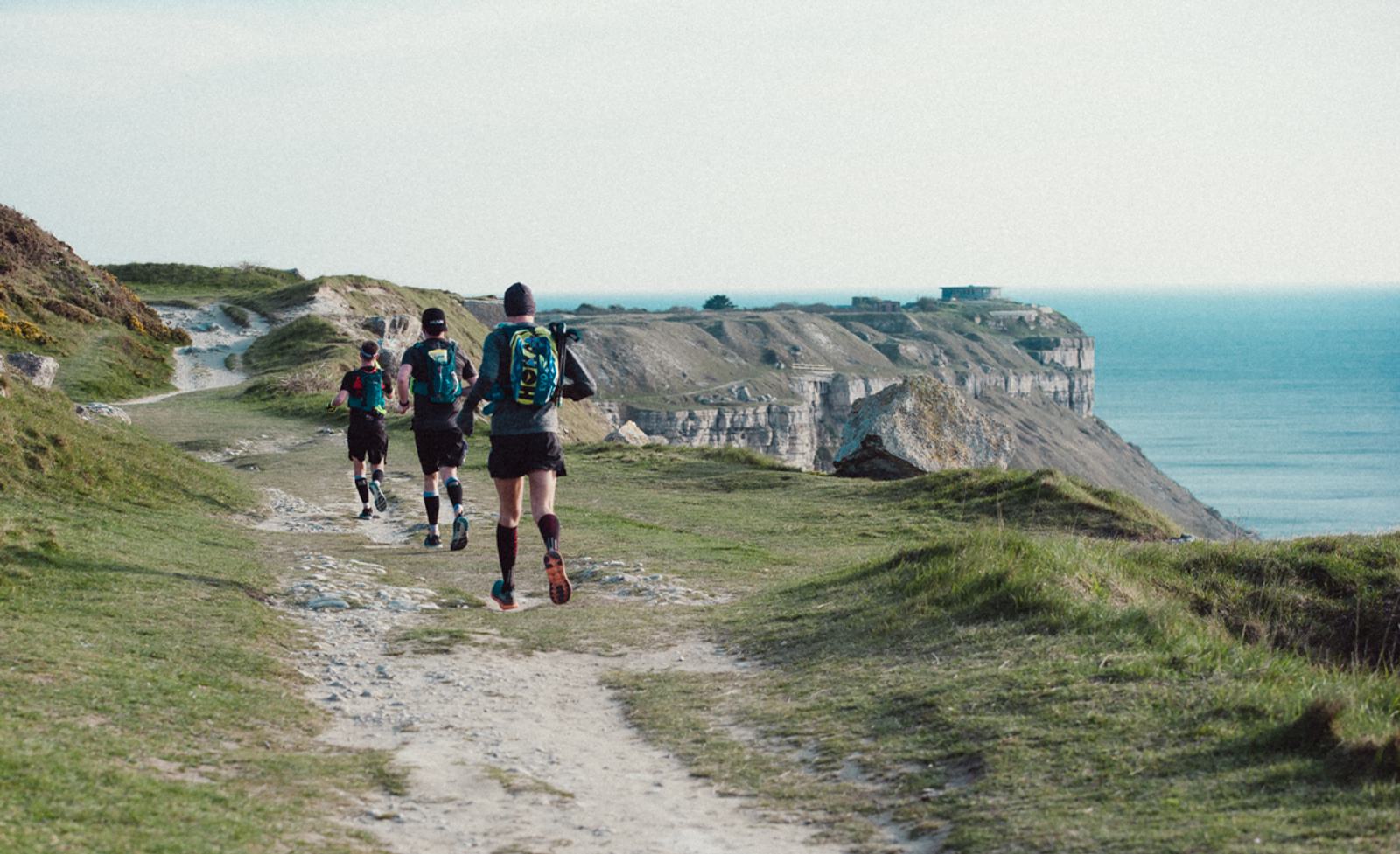 Three runners on a dirt trail along a grassy coastal cliff, with the ocean visible in the background. They wear athletic gear and backpacks, moving along a scenic path under a clear sky.