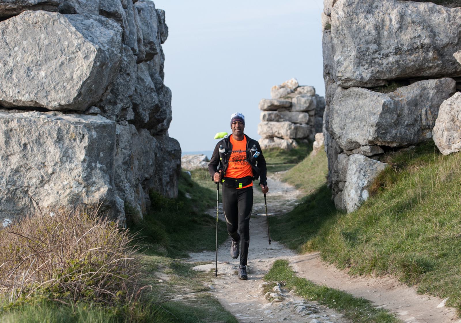 A trail runner wearing an orange shirt and black pants jogs with trekking poles on a rocky path between large stone formations. The sky is clear, and grass borders the trail.