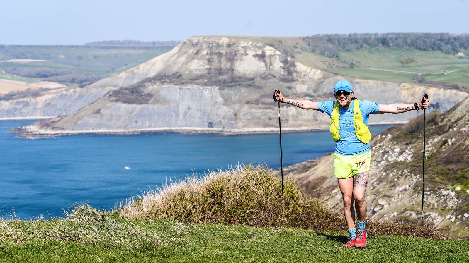 A person in bright athletic clothing and a cap is hiking with poles on a grassy coastal trail. The background features cliffs and a blue ocean under a clear sky.