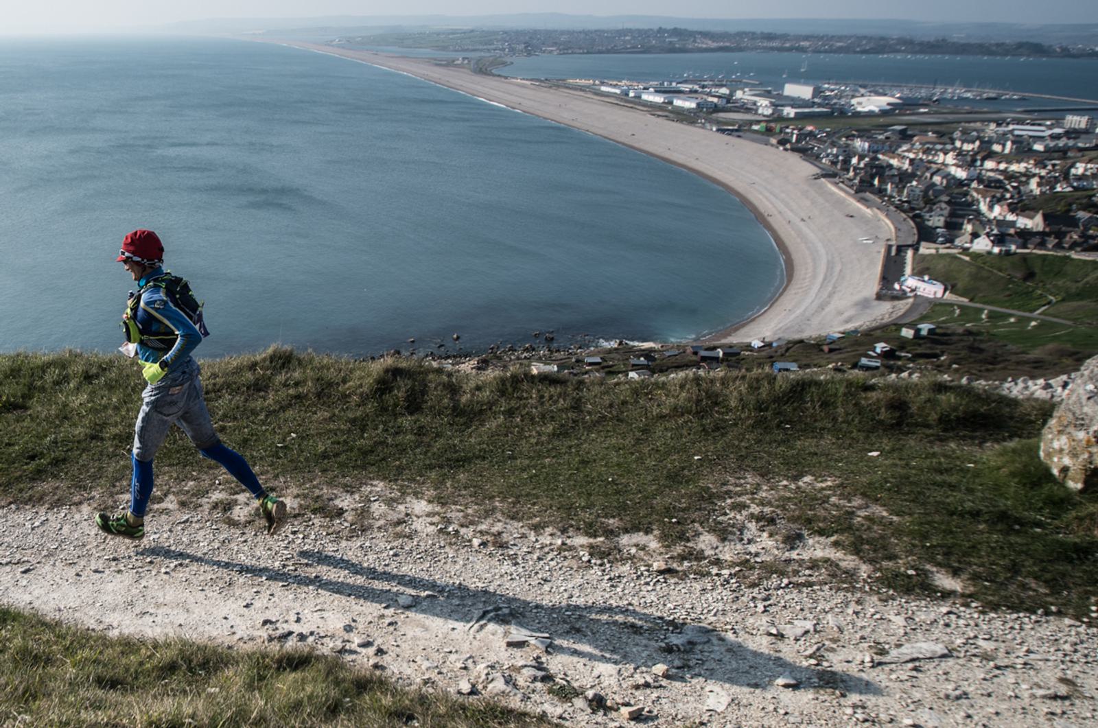 A runner in colorful sports gear jogs along a coastal path. The view includes a sweeping beach and the sea on the left, with a town in the background. The scene depicts a scenic, sunny day.