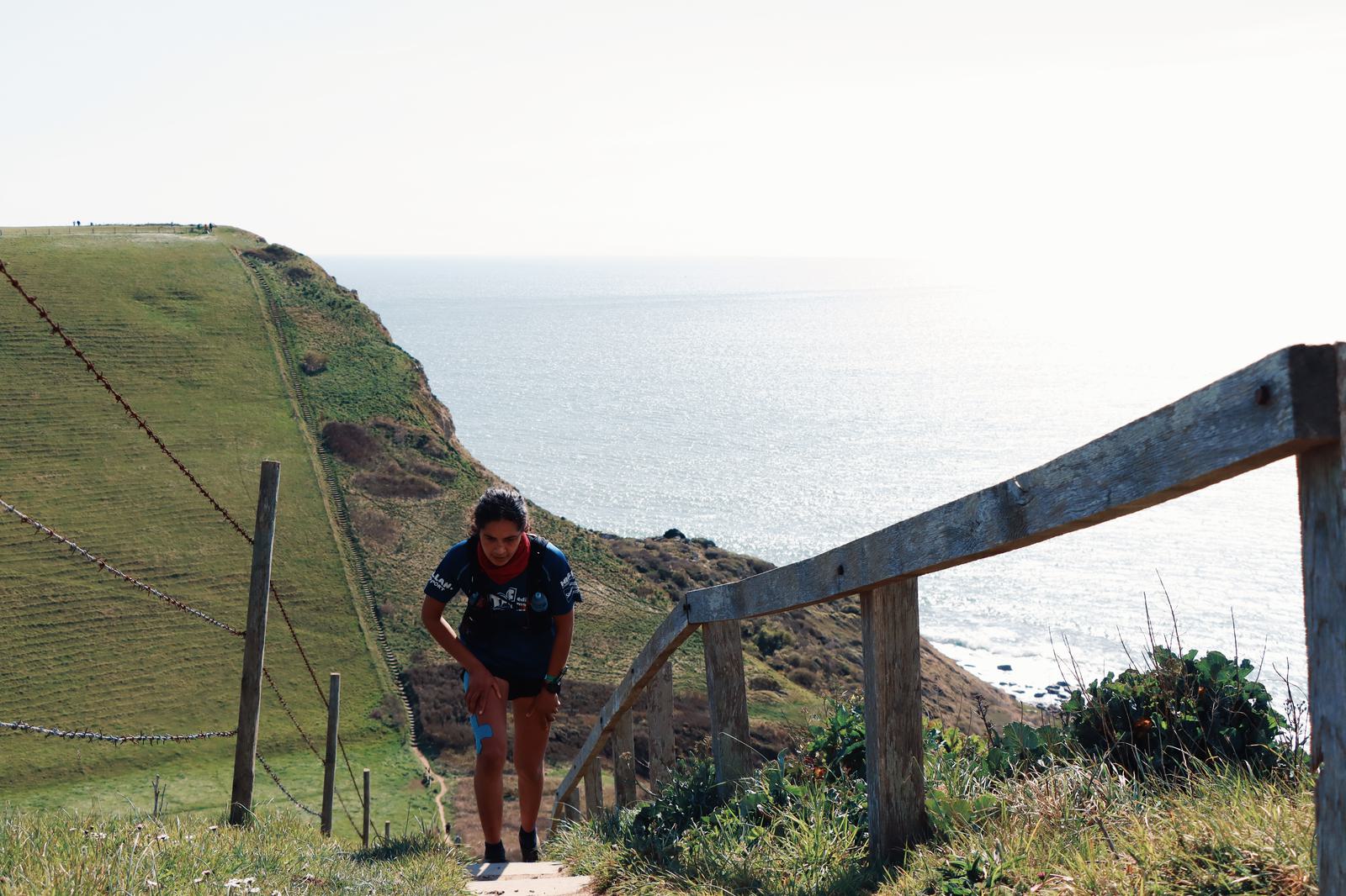 A person is walking up a steep path beside a wooden fence on a grassy hillside overlooking the ocean on a sunny day. The person appears to be slightly bent forward, with a scenic view of the sea and grassy cliffs in the background.
