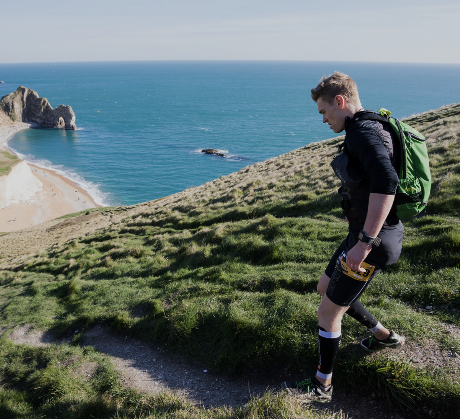 A person wearing a backpack hikes up a grassy hillside overlooking a turquoise sea and a sandy beach with distant rock formations on a sunny day.