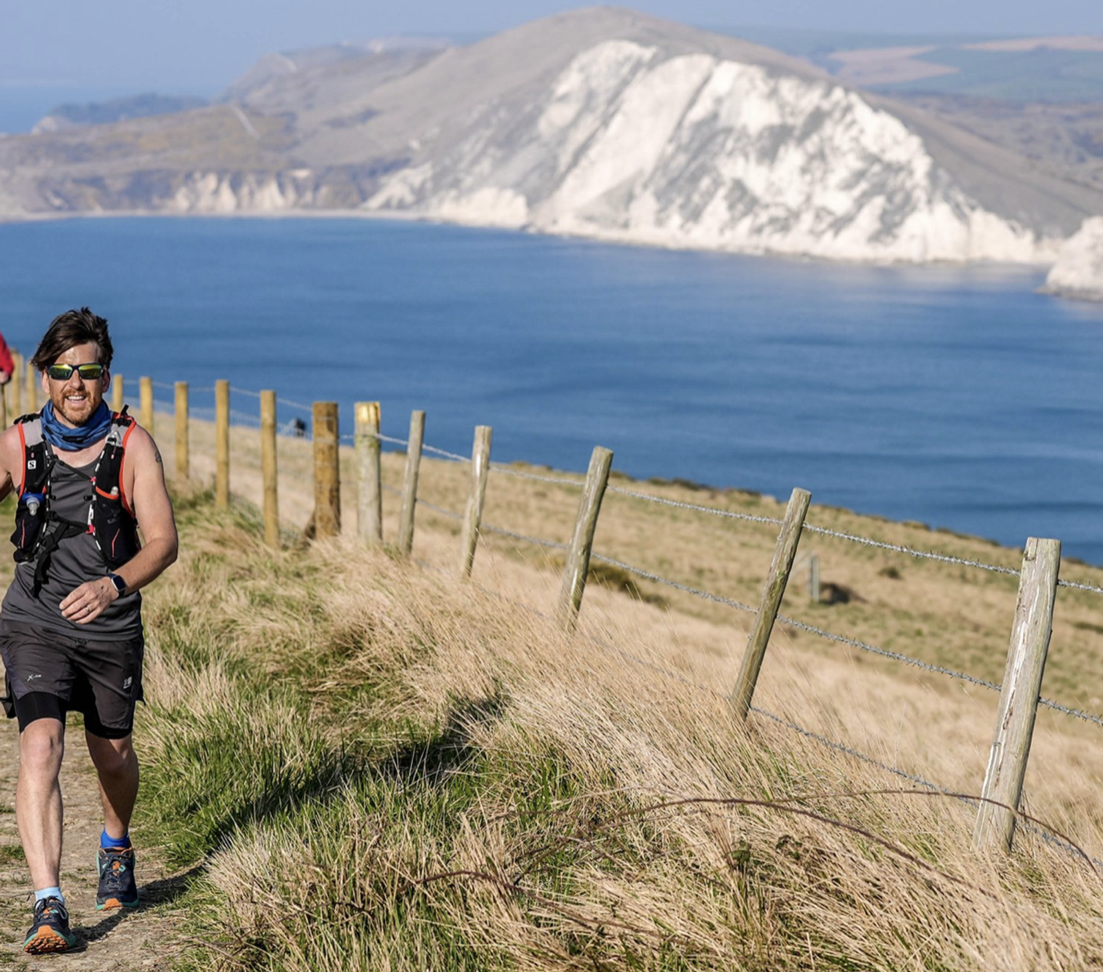 A person in athletic gear is running along a grassy seaside path, with a wooden fence on the right. The background features a calm blue ocean and white cliffs under a clear sky.