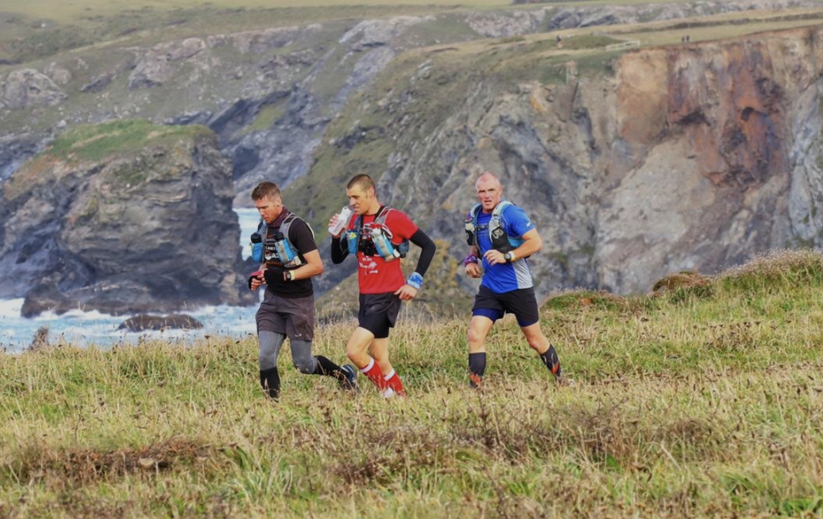 Three runners on a grassy trail near a cliff with a rocky coastline in the background. They wear running gear, with the ocean and rugged cliffs visible behind them under a cloudy sky.
