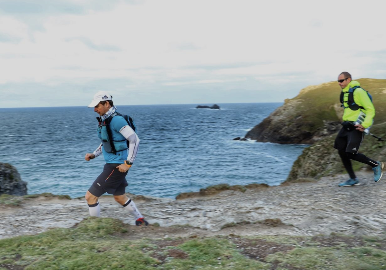 Two people are trail running along a coastal path. The ocean is visible on the left, and grassy cliffs are on the right. One person wears a white hat and blue jacket, the other wears a yellow jacket. The sky is partly cloudy.
