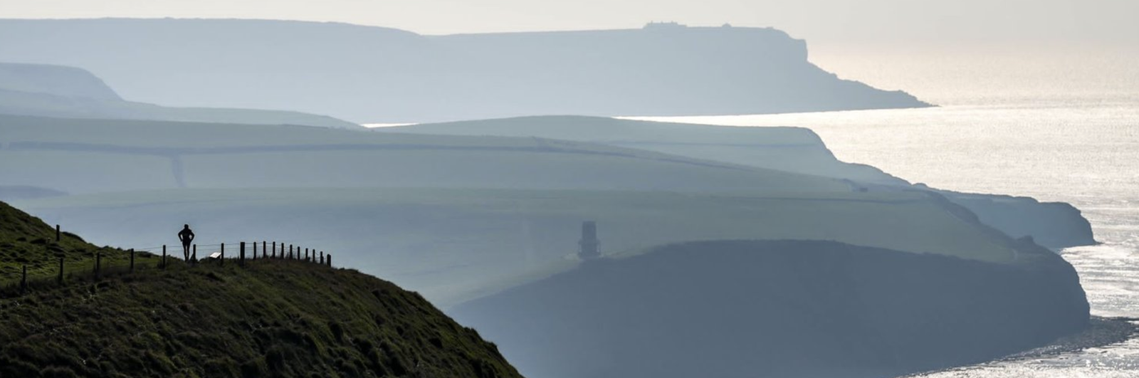 A lone cyclist rides along a grassy coastal trail with a distant view of layered cliffs and the sea under an overcast sky. The landscape is bathed in muted light, creating a serene and expansive atmosphere.