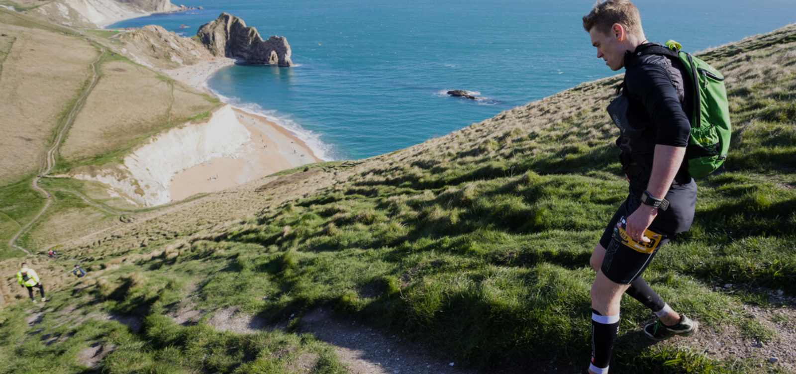 A person with a green backpack hikes uphill on a grassy trail, overlooking a scenic coastal view with cliffs, turquoise water, and a sandy beach below on a sunny day.