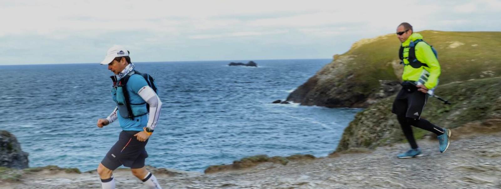 Two men wearing running gear jog along a rocky coastal path. The ocean is visible in the background, with waves crashing against the cliffs under an overcast sky. One runner wears a blue vest, while the other is in a neon green jacket.