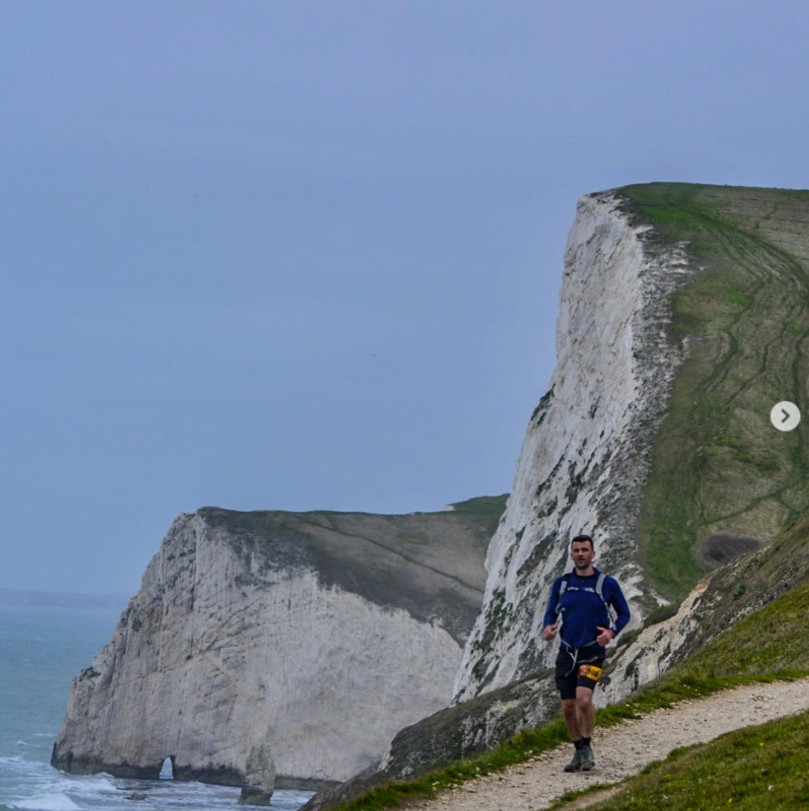 A person jogs along a cliffside path with dramatic white cliffs and the sea in the background. The scene is overcast, creating a moody atmosphere.