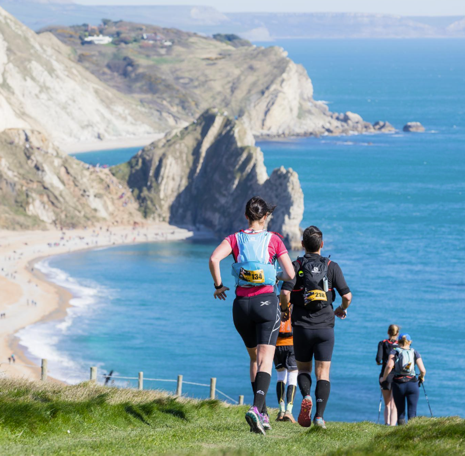 Two people run along a grassy cliff path overlooking a scenic coastline with sandy beaches and rugged cliffs. The ocean is bright blue under a clear sky, and several other runners are visible ahead.