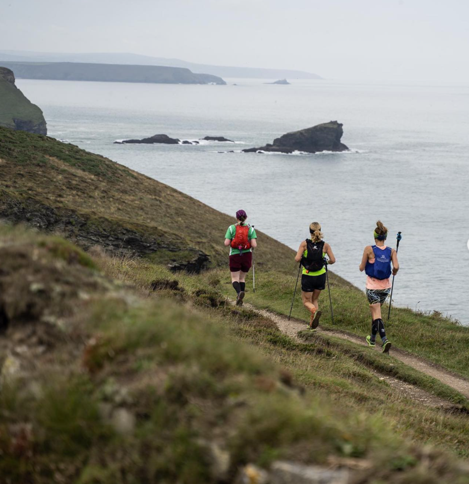 Three people trail running along a coastal path. They are equipped with backpacks and trekking poles. The path follows a grassy hillside, with a view of the ocean and rocky islands in the background. The sky is overcast.